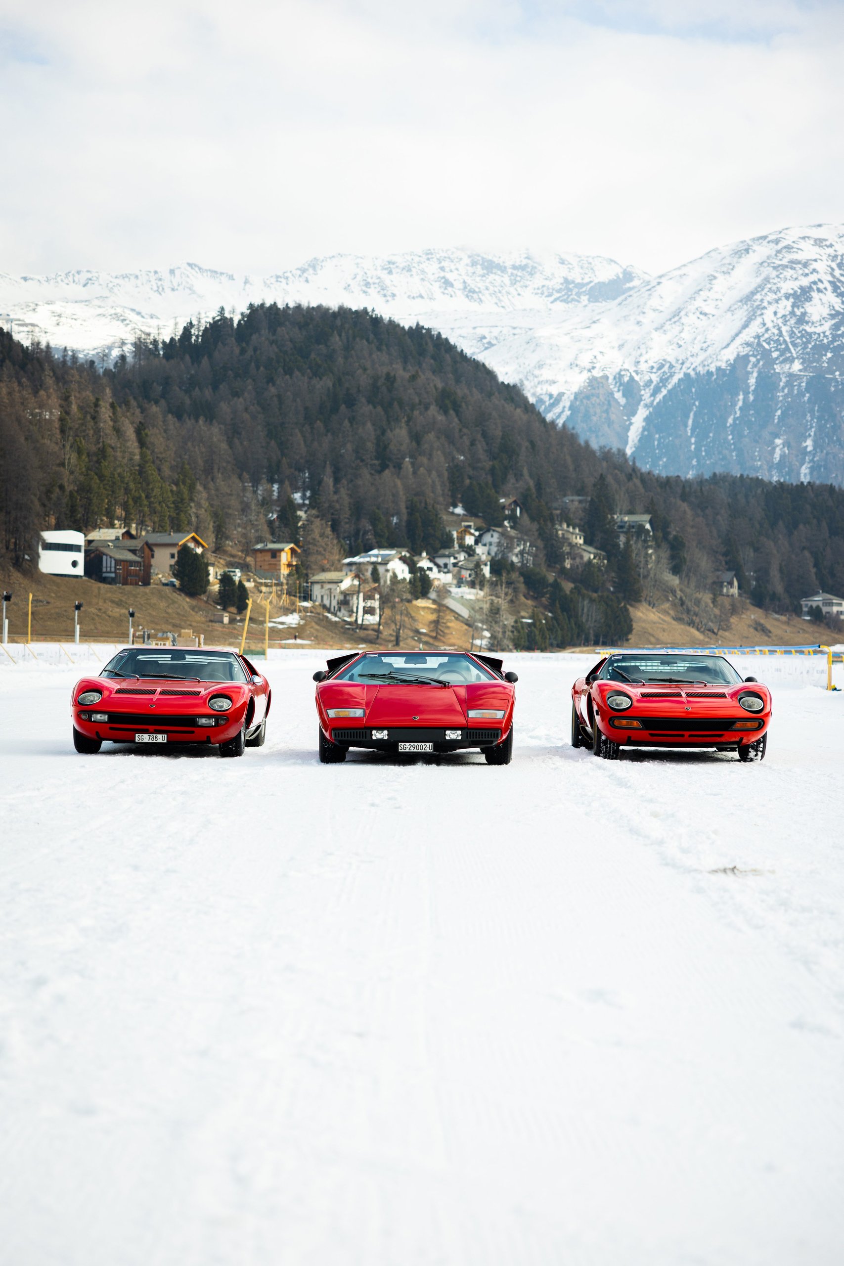 Three red classic lamborghinis lined up on snow at st. Moritz: miura sv, countach lp400, and miura p400s with mountains in background