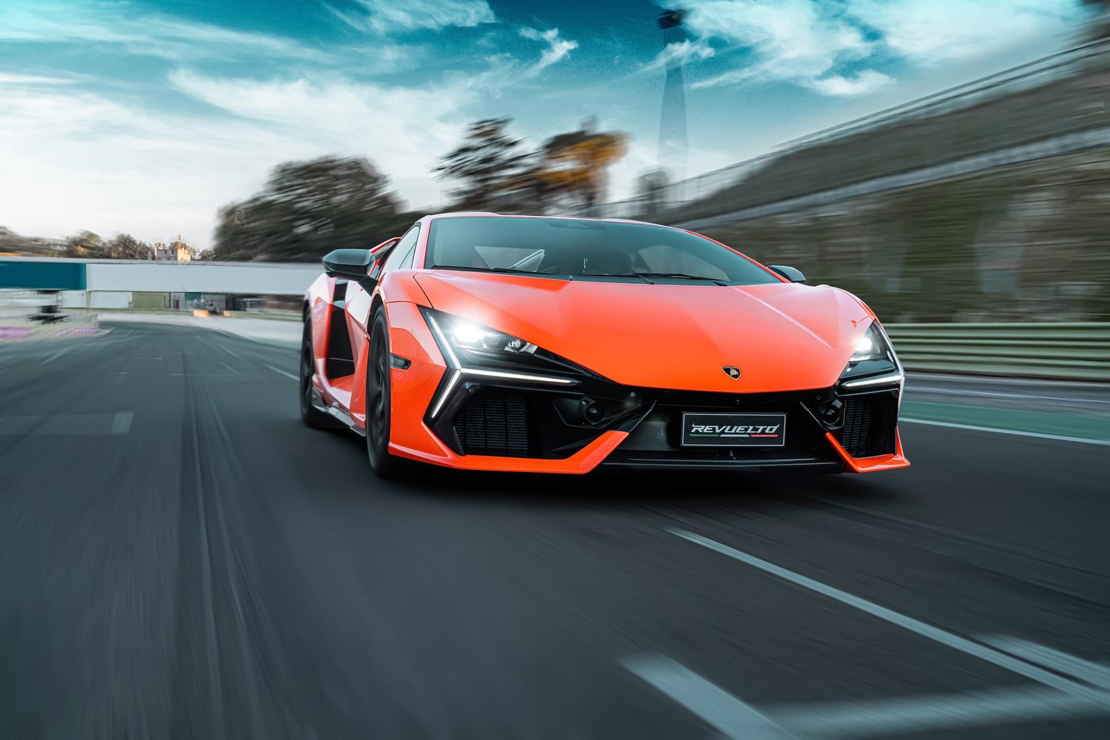 Orange Lamborghini Revuelto at speed on the Vallelunga circuit, Y-shaped DRLs and front splitter visible in motion blur