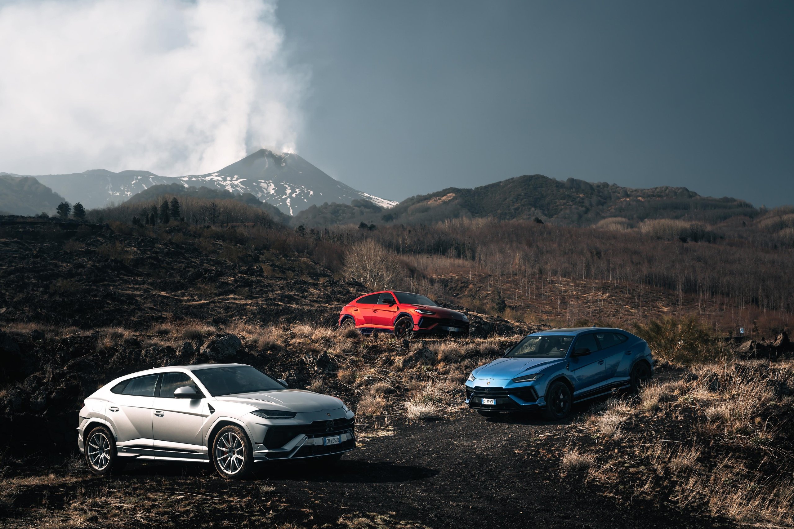 Three Lamborghini Urus SUVs in silver, red, and blue parked on a volcanic landscape