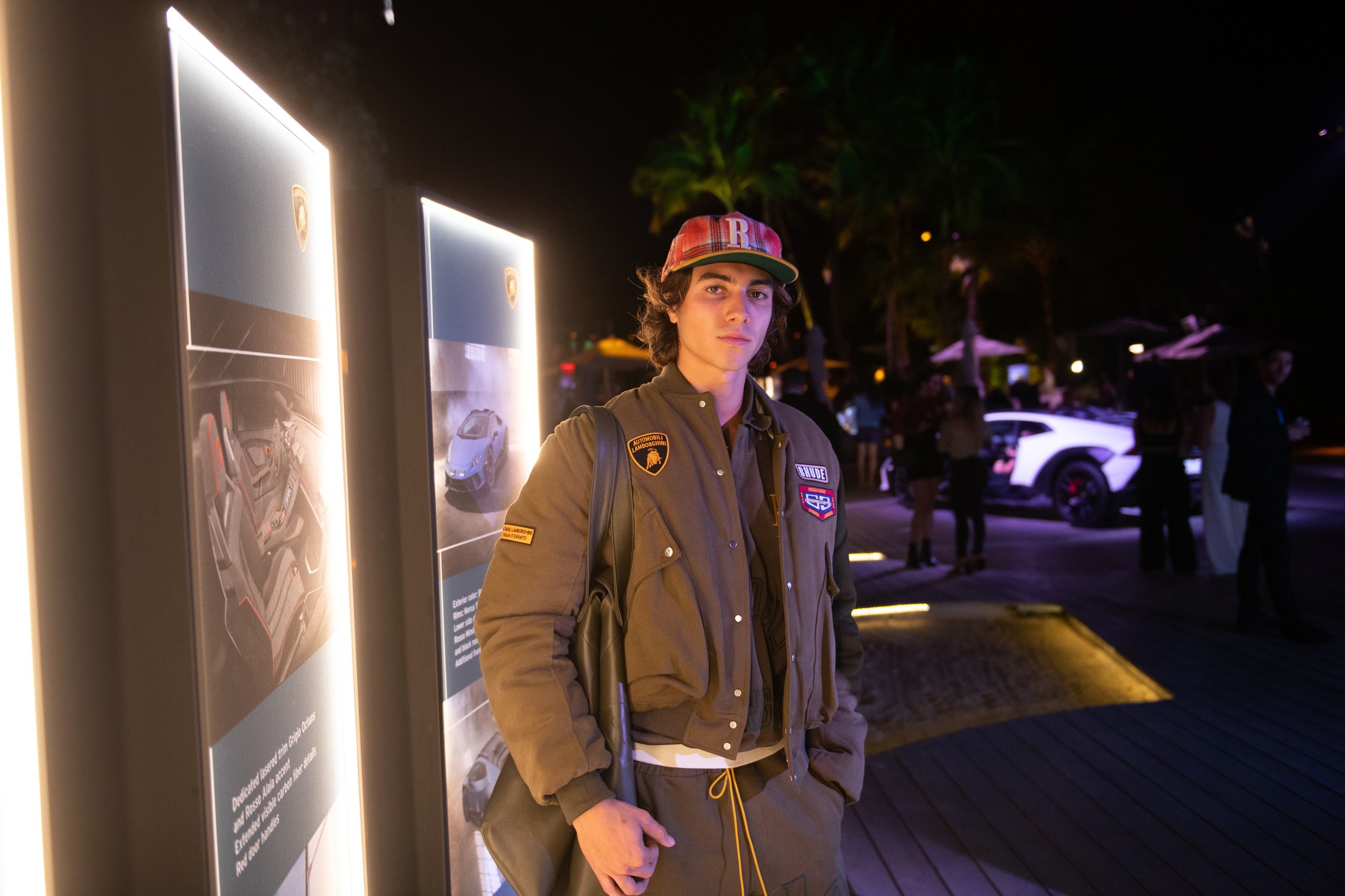 Rhude X Automobili Lamborghini: A Sterrato-Inspired Capsule That Reveals Where The Brand Is Headed 3 A model wearing a green rhude x lamborghini jacket and cap at the art basel miami evening event, with a white lamborghini visible in the background