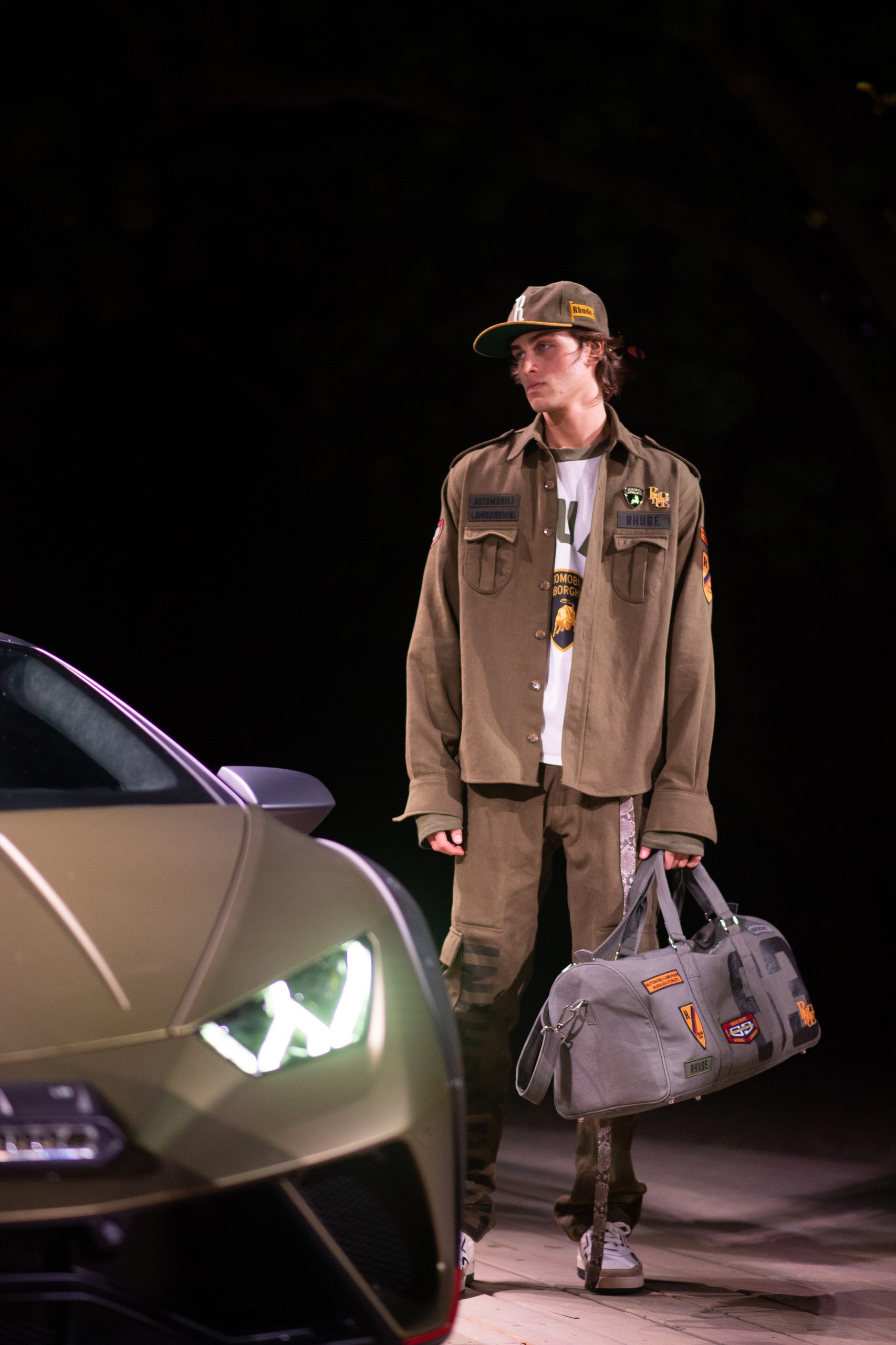 Rhude X Automobili Lamborghini: A Sterrato-Inspired Capsule That Reveals Where The Brand Is Headed 7 A model in rhude x lamborghini military-style apparel stands beside a gold huracán sterrato, holding a branded canvas duffel bag