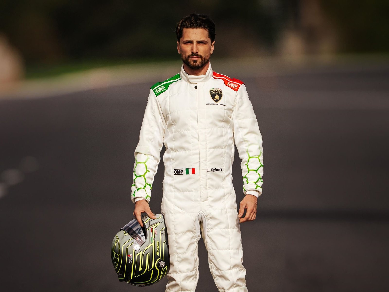 Loris Spinelli standing on a race track in his Lamborghini Squadra Corse racing suit, holding his helmet