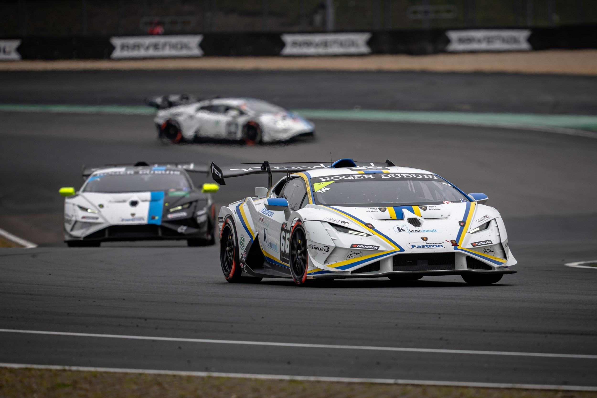 A white lamborghini huracán super trofeo evo2 leads two other race cars through a corner showcasing close competitive action