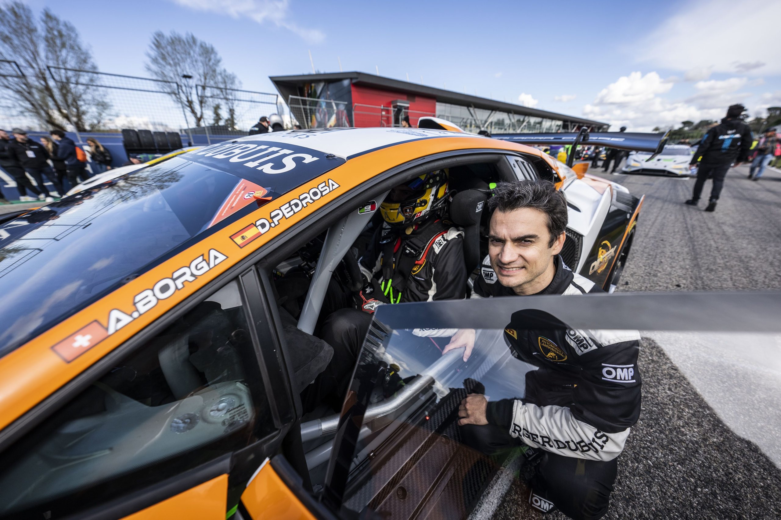 Pedrosa Returns, A Perfect Streak Faces Misano'S Evening Grid, And Four Classes Fight For Points 3 A man in racing attire smiling while leaning out of a white and orange lamborghini huracán super trofeo evo2, with a driver visible inside the cockpit