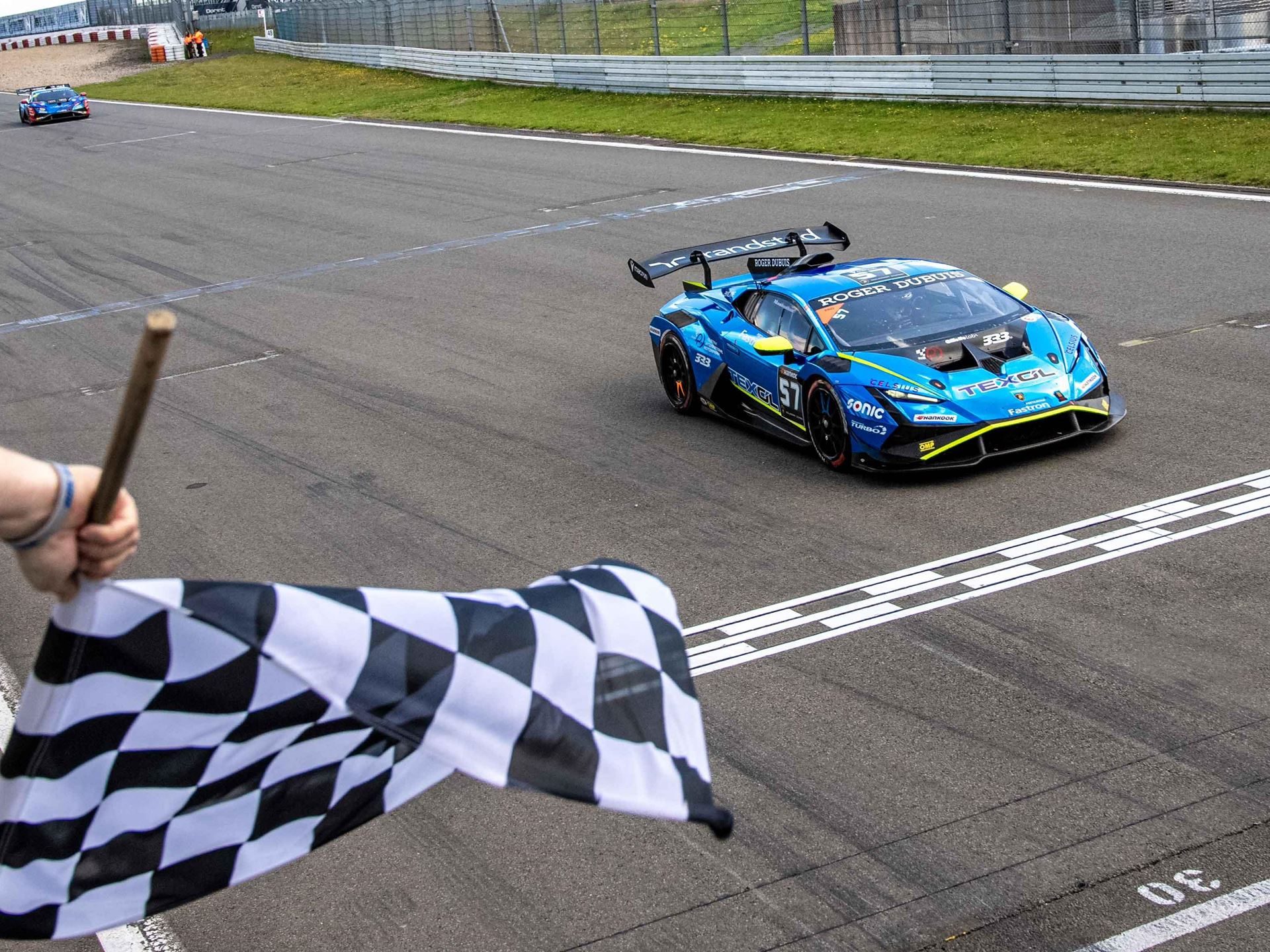 Split Weather, Split Winners: Bonduel And Orudzhev Trade Nürburgring Victories In Super Trofeo Europe 5 A blue and yellow lamborghini huracán super trofeo evo crosses the finish line as a checkered flag is waved
