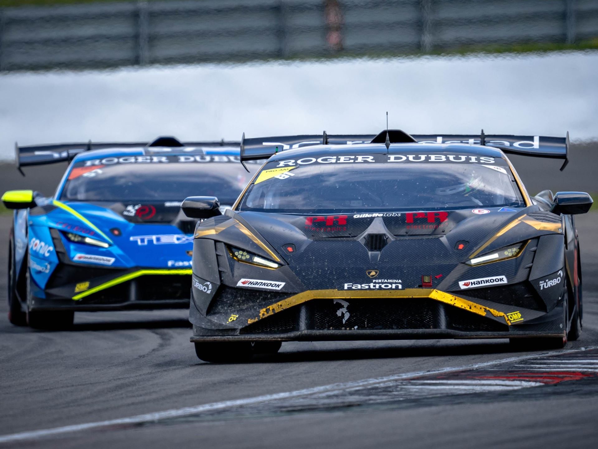 Split Weather, Split Winners: Bonduel And Orudzhev Trade Nürburgring Victories In Super Trofeo Europe 7 A black and gold lamborghini huracán super trofeo evo closely pursued by a blue and yellow rival on a dry nürburgring racetrack