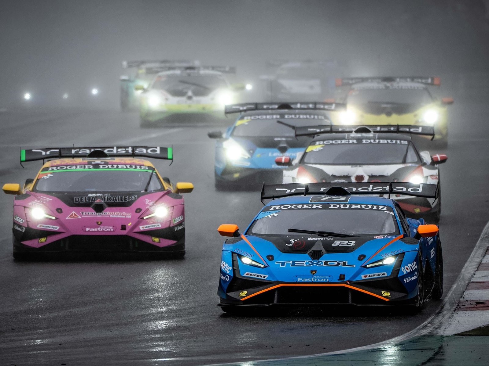 A blue and orange Lamborghini Huracán Super Trofeo EVO leads a pack of race cars on a wet Nürburgring track with headlights cutting through the mist