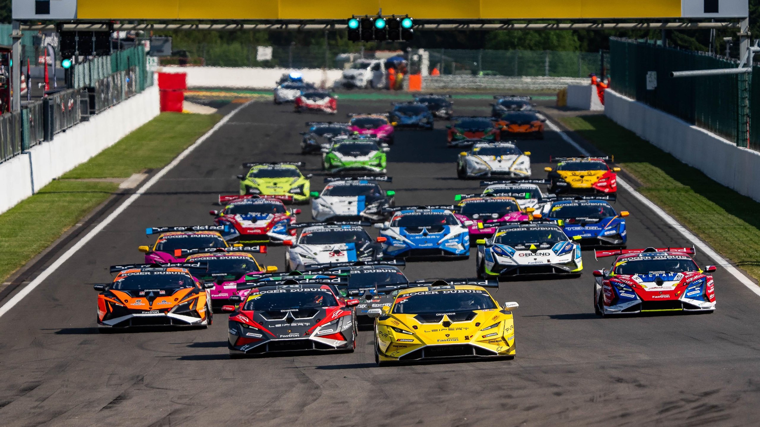 A large grid of lamborghini huracán super trofeo evo2 race cars lined up on the spa-francorchamps circuit under bright sunlight