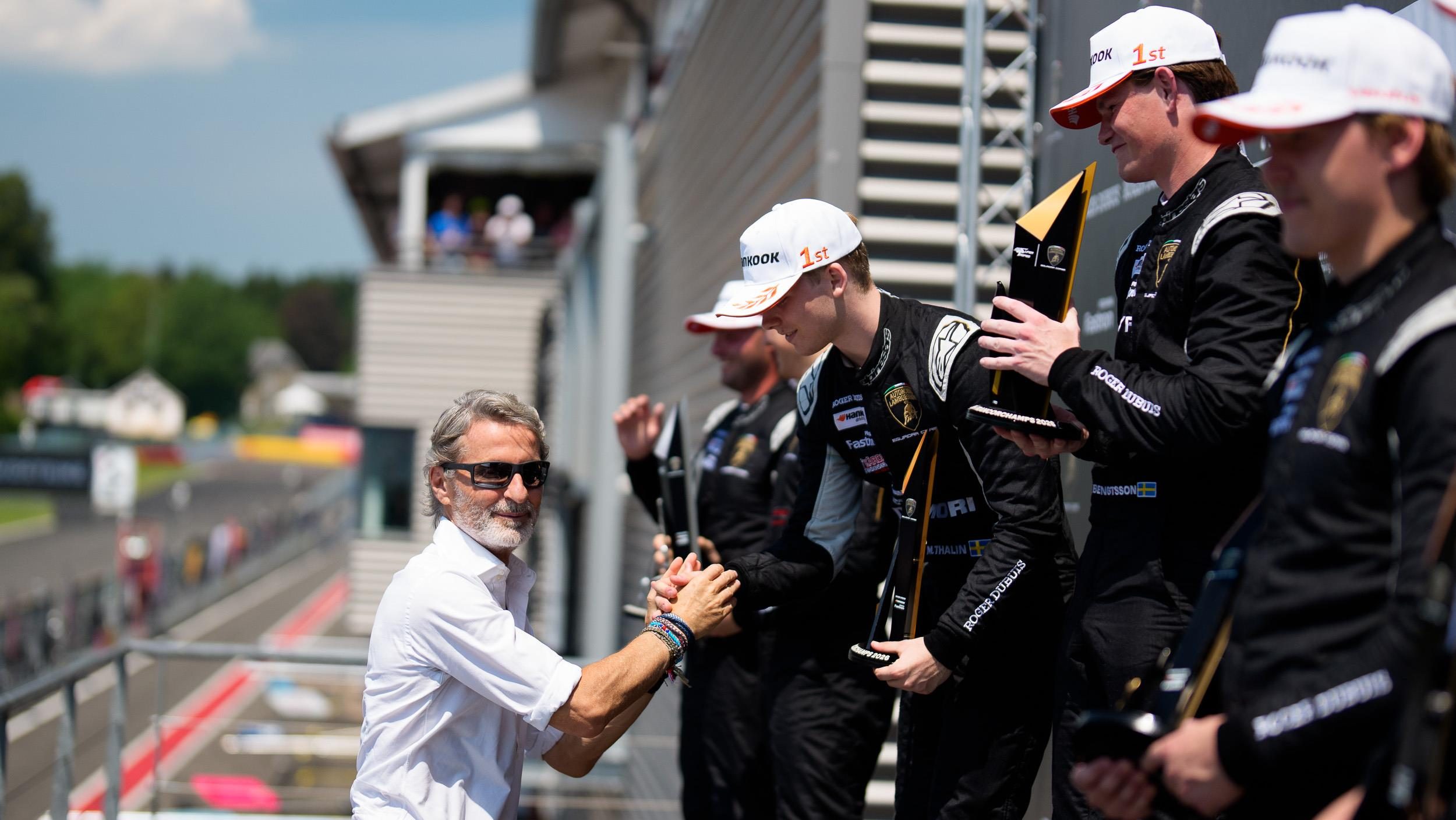 A podium ceremony at spa-francorchamps with a race official shaking hands with a winning super trofeo driver while teammates hold trophies