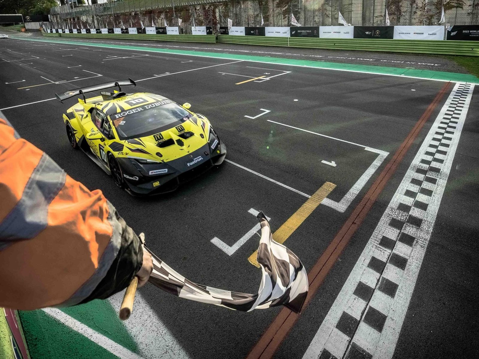 A yellow and black Lamborghini Huracán Super Trofeo EVO2 crosses the finish line under a checkered flag at Vallelunga