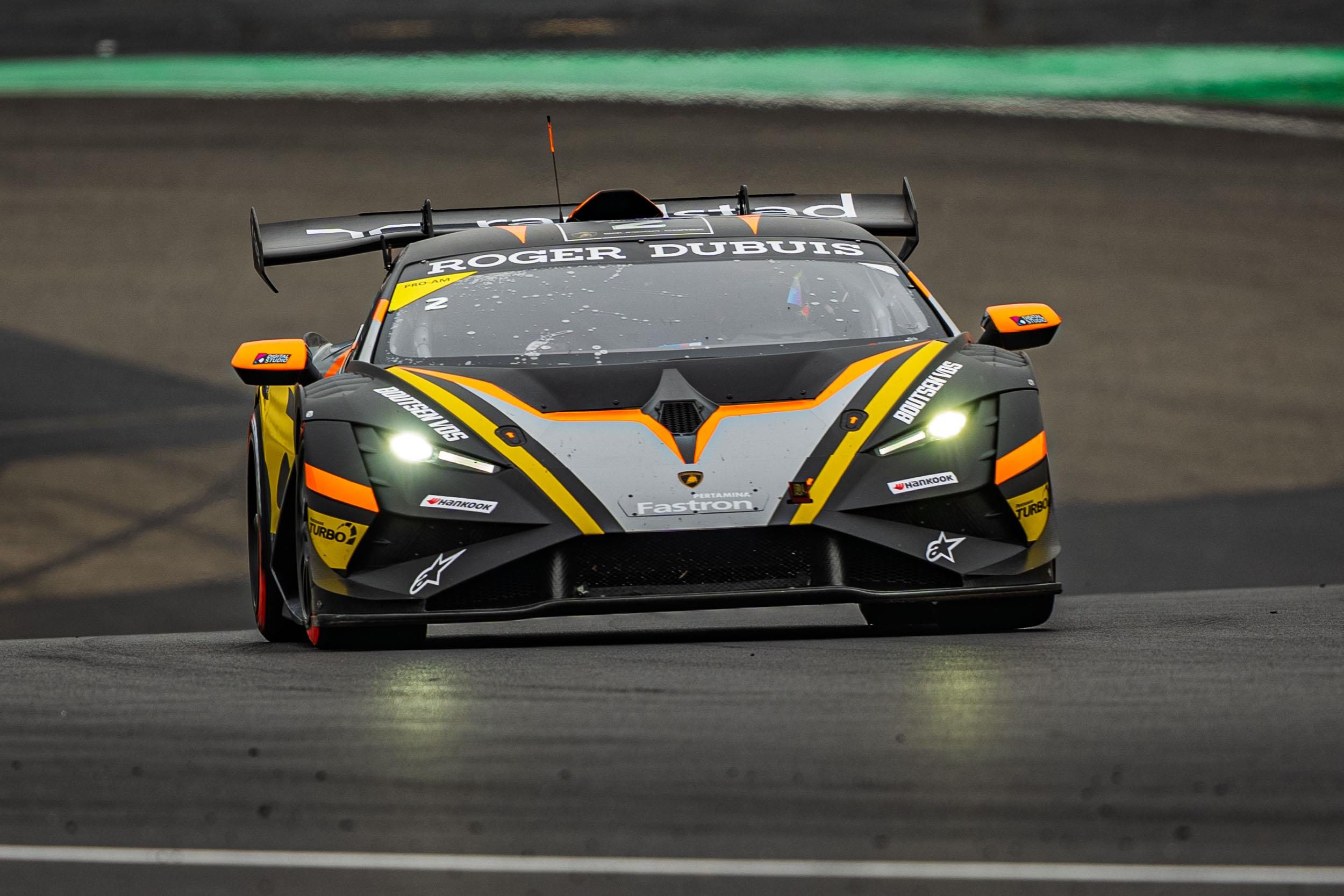 A black lamborghini huracán super trofeo evo2 with yellow and orange stripes racing with headlights on at the nürburgring