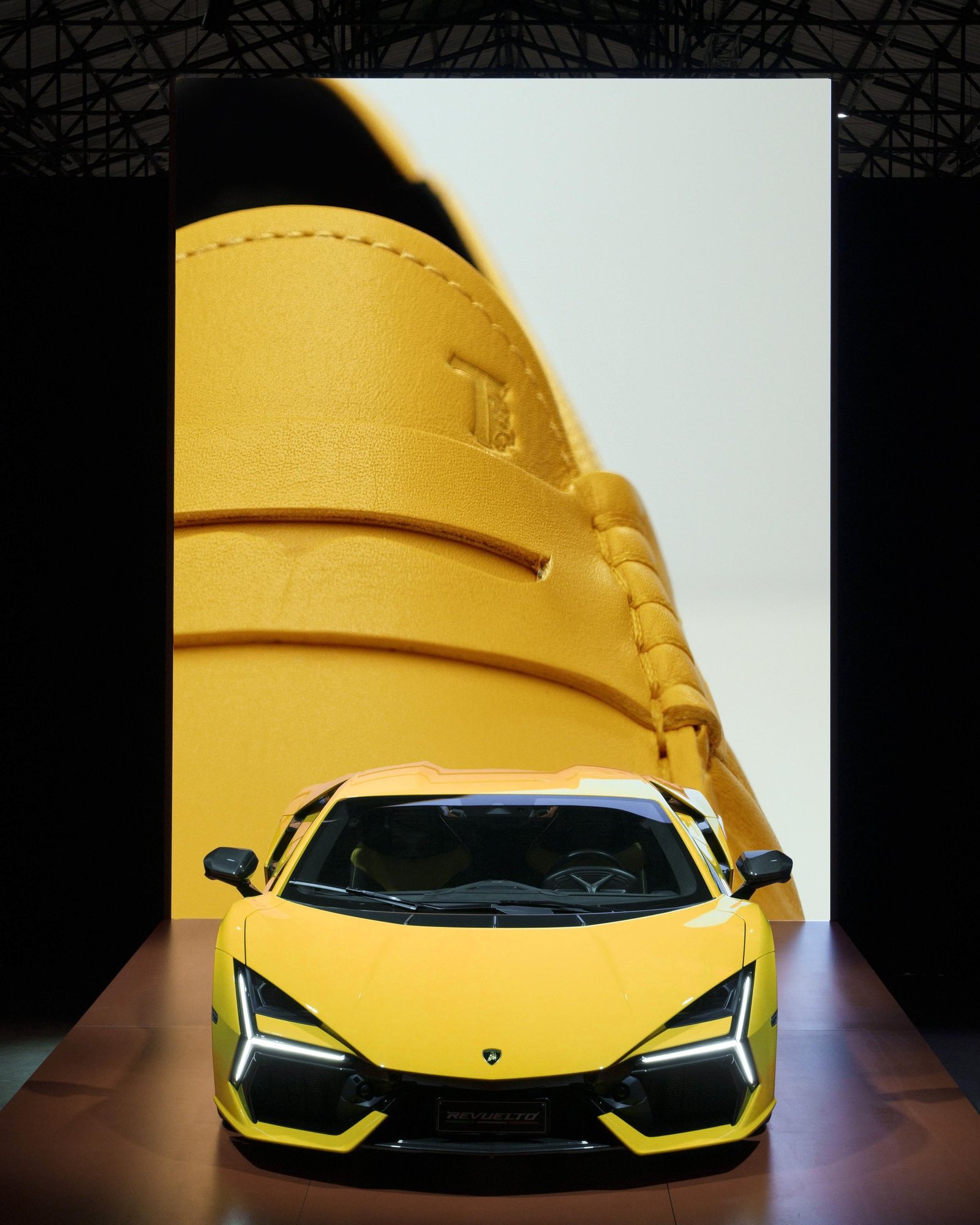 Yellow Lamborghini Revuelto displayed at the Tod's x Automobili Lamborghini launch event with a large screen showing a yellow Tod's loafer behind it