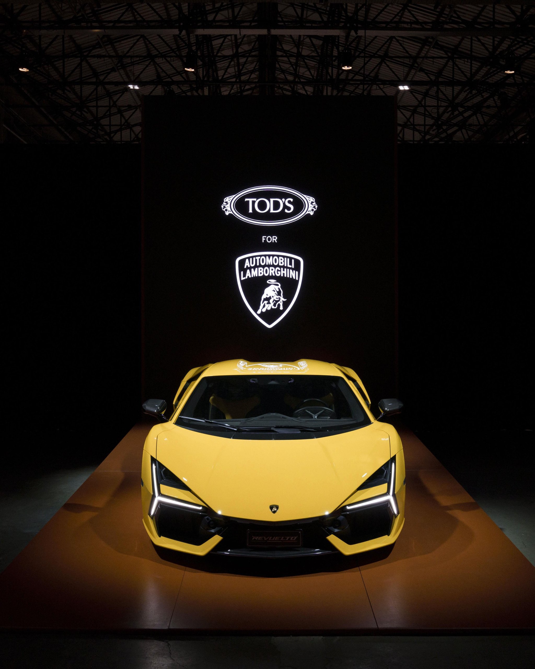 Tod'S X Automobili Lamborghini: What A Driving Shoe Tells You About Where The Brand Is Headed 3 Frontal view of a yellow lamborghini revuelto on a raised platform with tod's and automobili lamborghini logos illuminated on a black backdrop