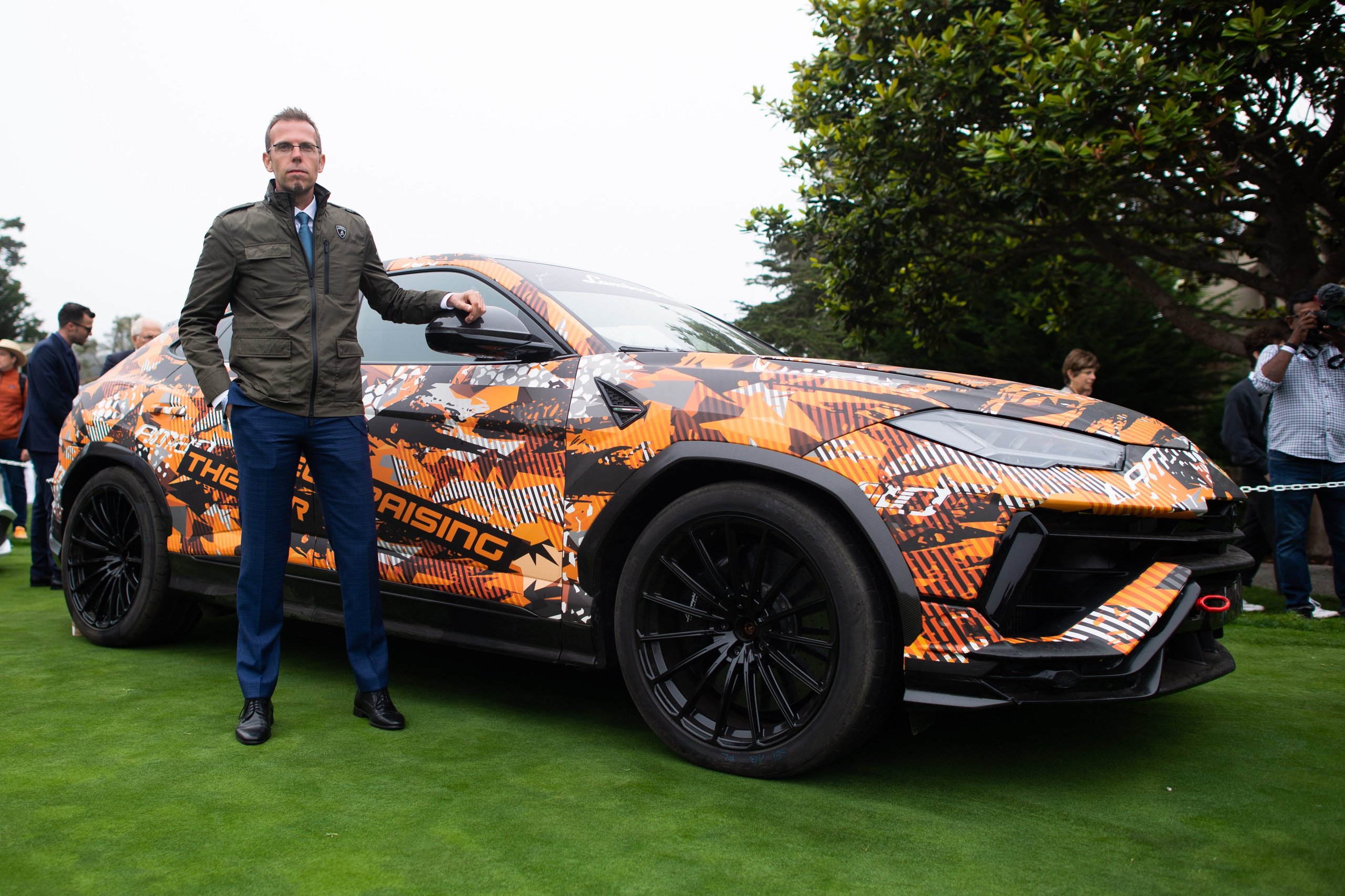 Lamborghini Brought A Mountain Record To The Most Exclusive Lawn In America 7 A man in a jacket and blue trousers stands next to the lamborghini urus performante in orange and black camouflage on the pebble beach lawn