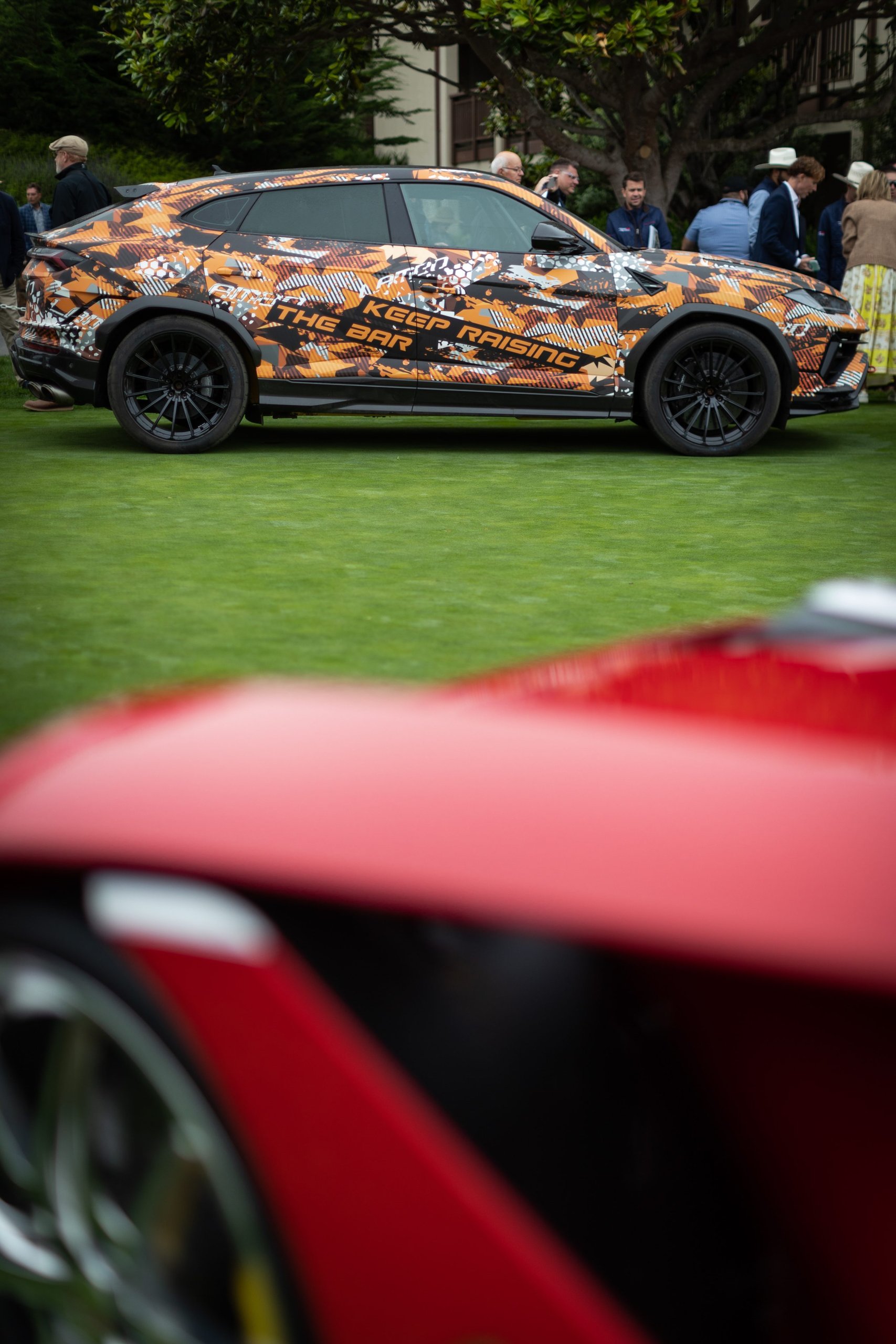 Lamborghini Brought A Mountain Record To The Most Exclusive Lawn In America 3 Lamborghini urus performante in orange and black camouflage livery parked on the green lawn at pebble beach, with a blurred red car in the foreground