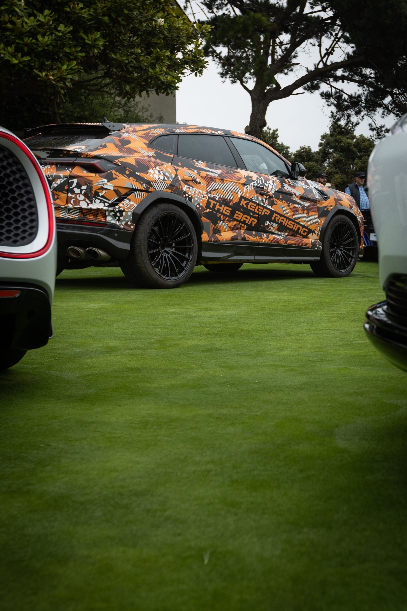 Lamborghini Brought A Mountain Record To The Most Exclusive Lawn In America 5 Rear three-quarter view of the lamborghini urus performante in orange and black camouflage on a green lawn, framed by blurred cars in the foreground
