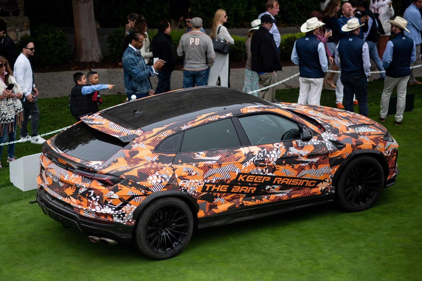 Lamborghini Urus Performante in orange and black camouflage livery displayed on the Concept Lawn at Pebble Beach Concours d'Elegance, surrounded by onlookers