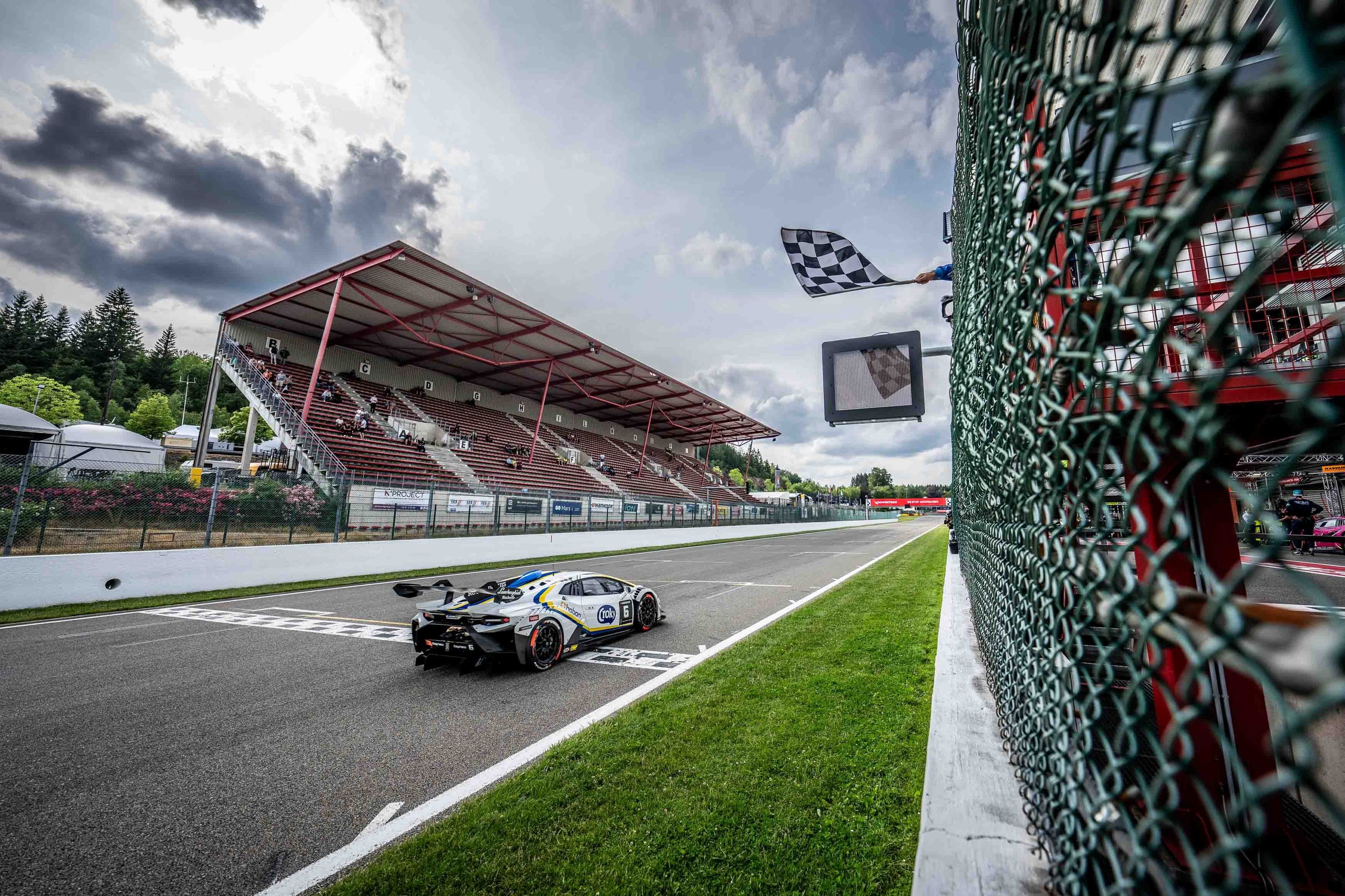 A lamborghini huracán super trofeo evo2 crosses the finish line with a checkered flag waving