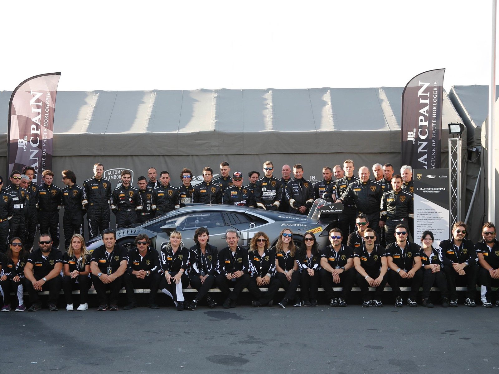 Lamborghini Squadra Corse team poses with a Huracan GT3 race car in front of sponsor banners and team tent