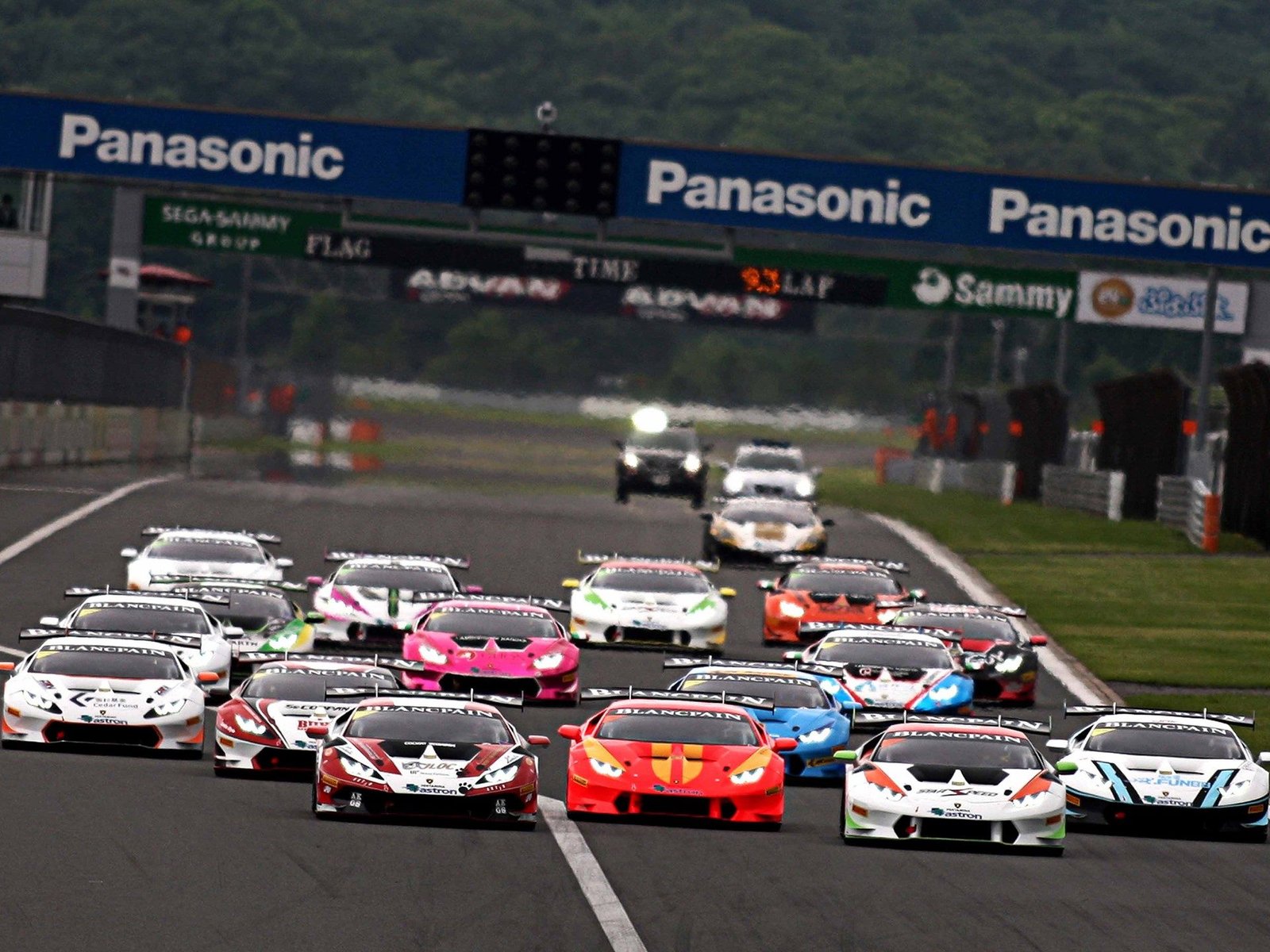 A pack of multi-colored Lamborghini Huracán GT3 race cars battling for position on a wet racetrack during the Blancpain Super Trofeo series