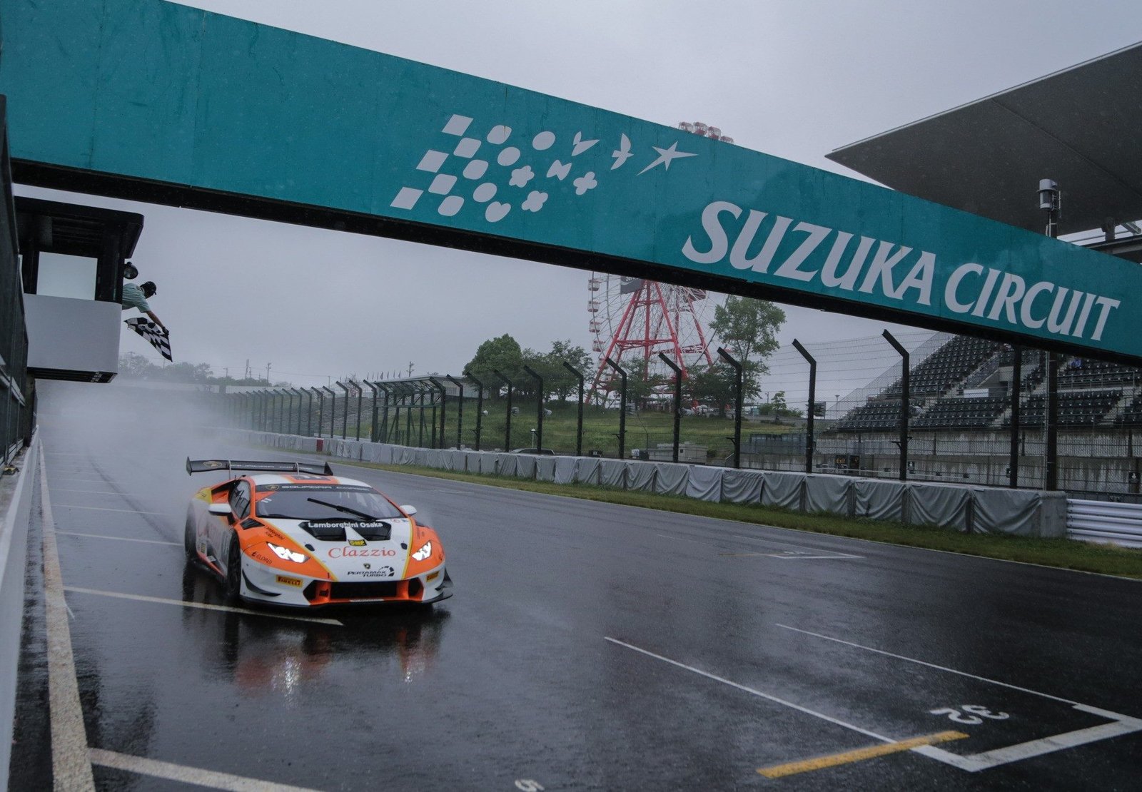 White and orange Lamborghini Huracán Super Trofeo race car crossing under the Suzuka Circuit banner on a soaking wet track with a checkered flag waving in the background
