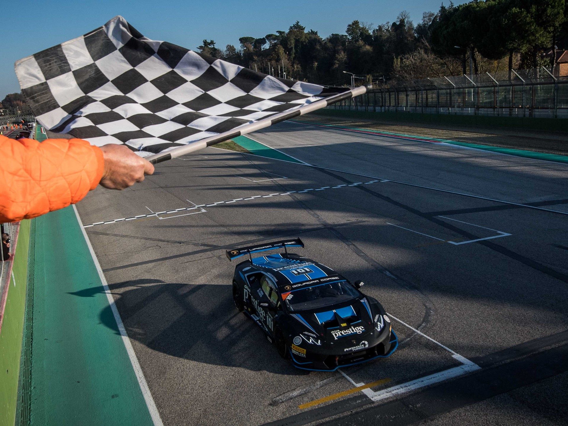 A black Lamborghini Huracan Super Trofeo race car with blue accents crosses the finish line under a waving checkered flag at Imola