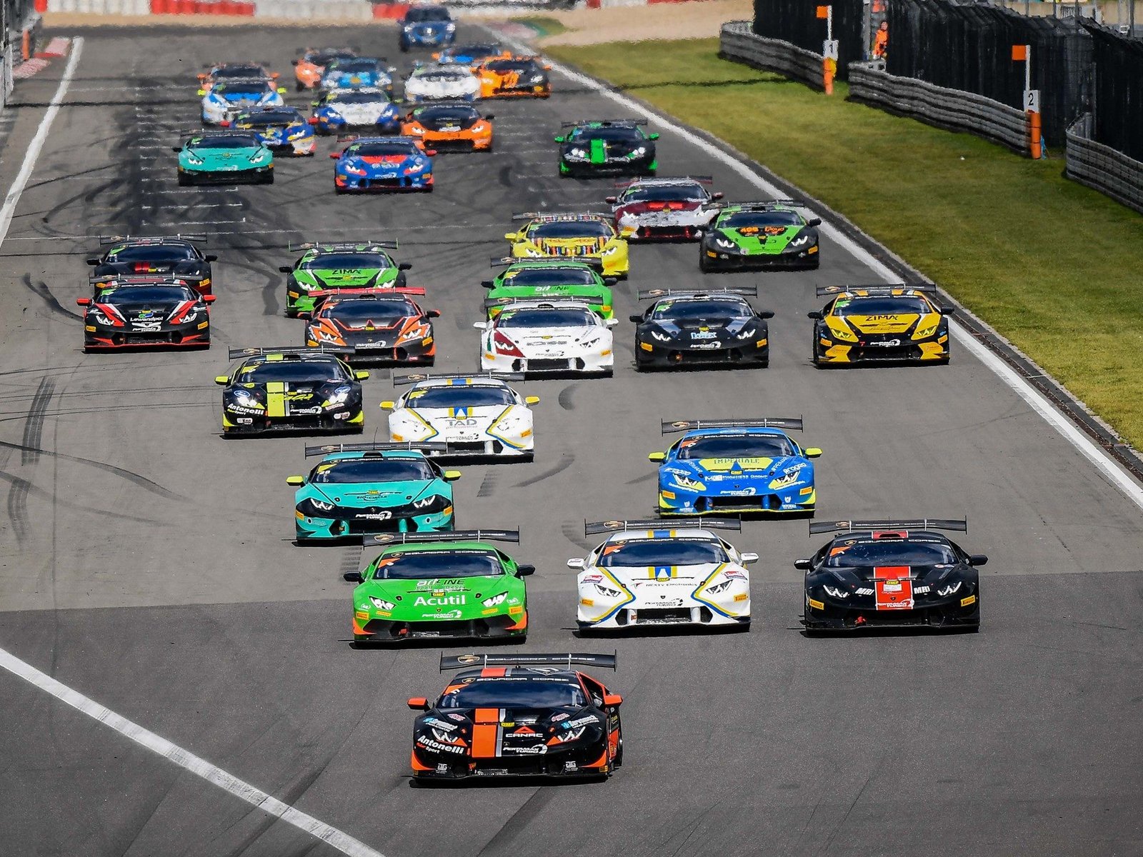 Large field of multi-colored Lamborghini Huracán Super Trofeo race cars battling for position on a racetrack under bright daylight