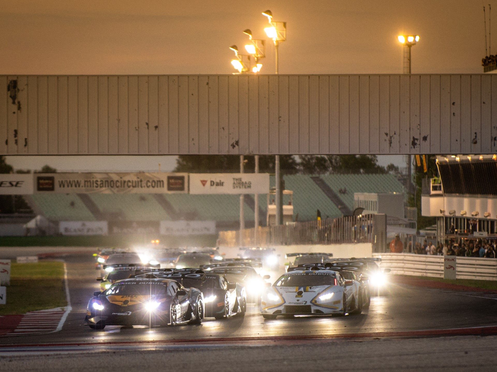 Lamborghini Huracán Super Trofeo EVO race cars with headlights on racing in a tight pack under stadium lights at Misano circuit
