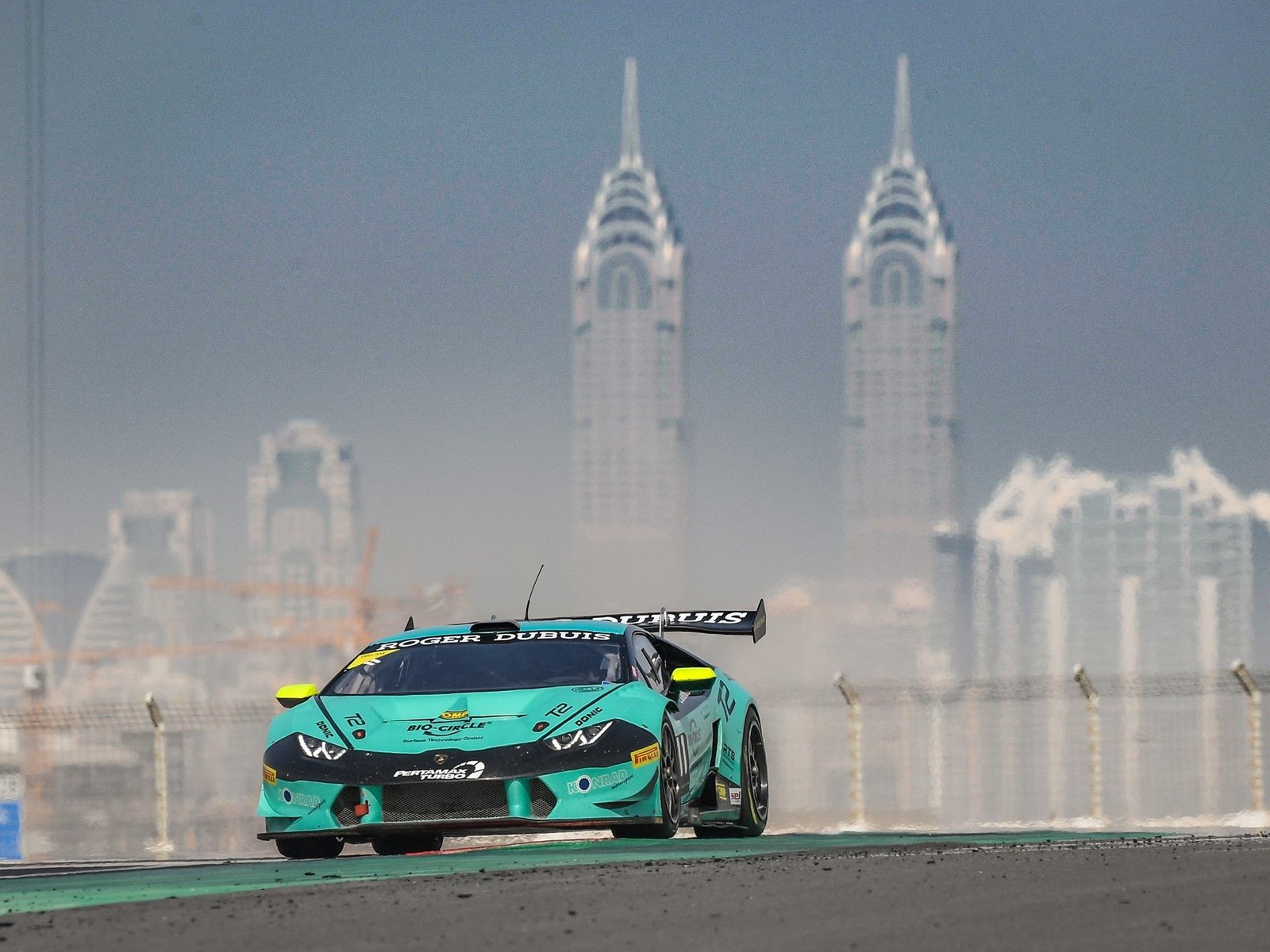 Teal and black Lamborghini Huracán Super Trofeo Evo race car at speed on track with city skyline in background