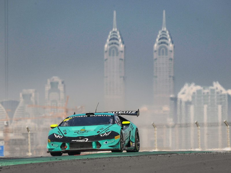 Teal and black Lamborghini Huracán Super Trofeo Evo race car at speed on track with city skyline in background