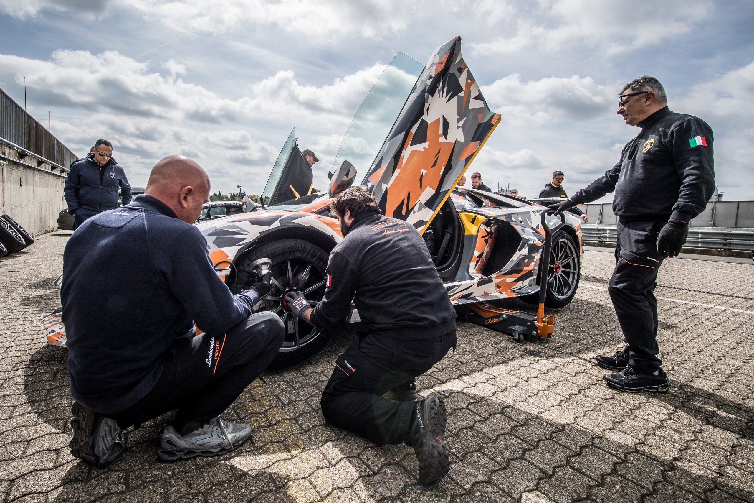 Camouflaged Aventador SVJ with scissor doors open, mechanics working on wheels in pit garage at the Nürburgring