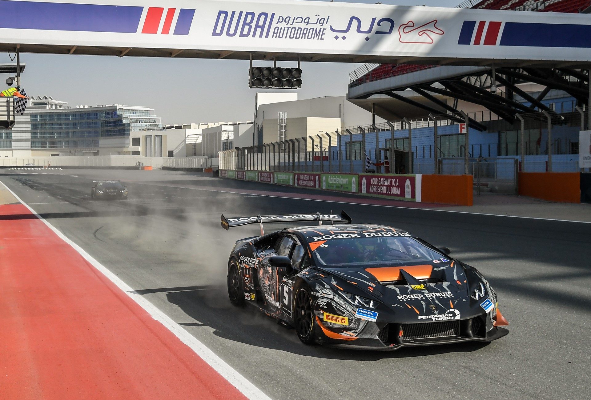 Black and orange Lamborghini Huracán race car at speed on the Dubai Autodrome circuit, kicking up dust with another competitor visible in the background