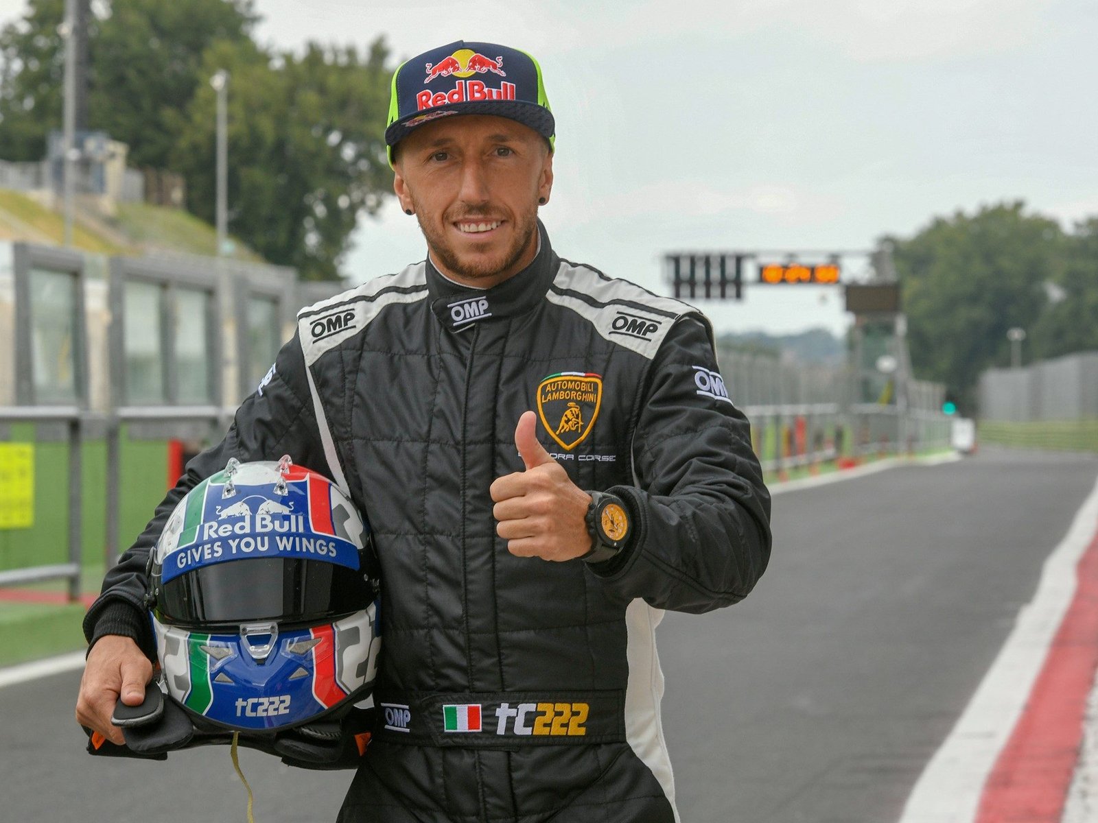 A race car driver in Lamborghini Squadra Corse racing suit gives a thumbs-up while holding a racing helmet on a circuit pit lane