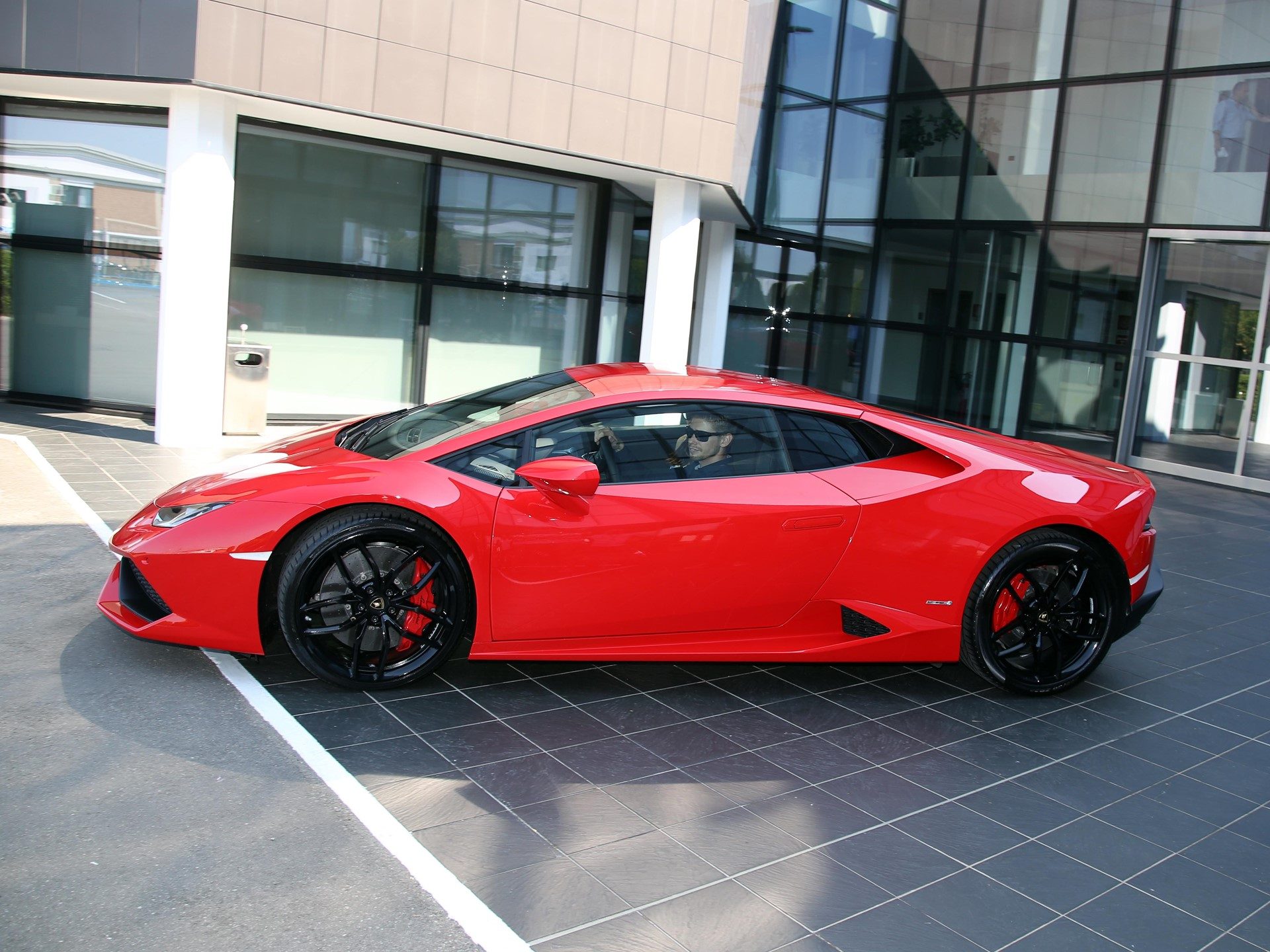 Red Lamborghini Huracán LP 610-4 with visible driver, parked on tiled surface showing sleek side profile and black wheels with red brake calipers