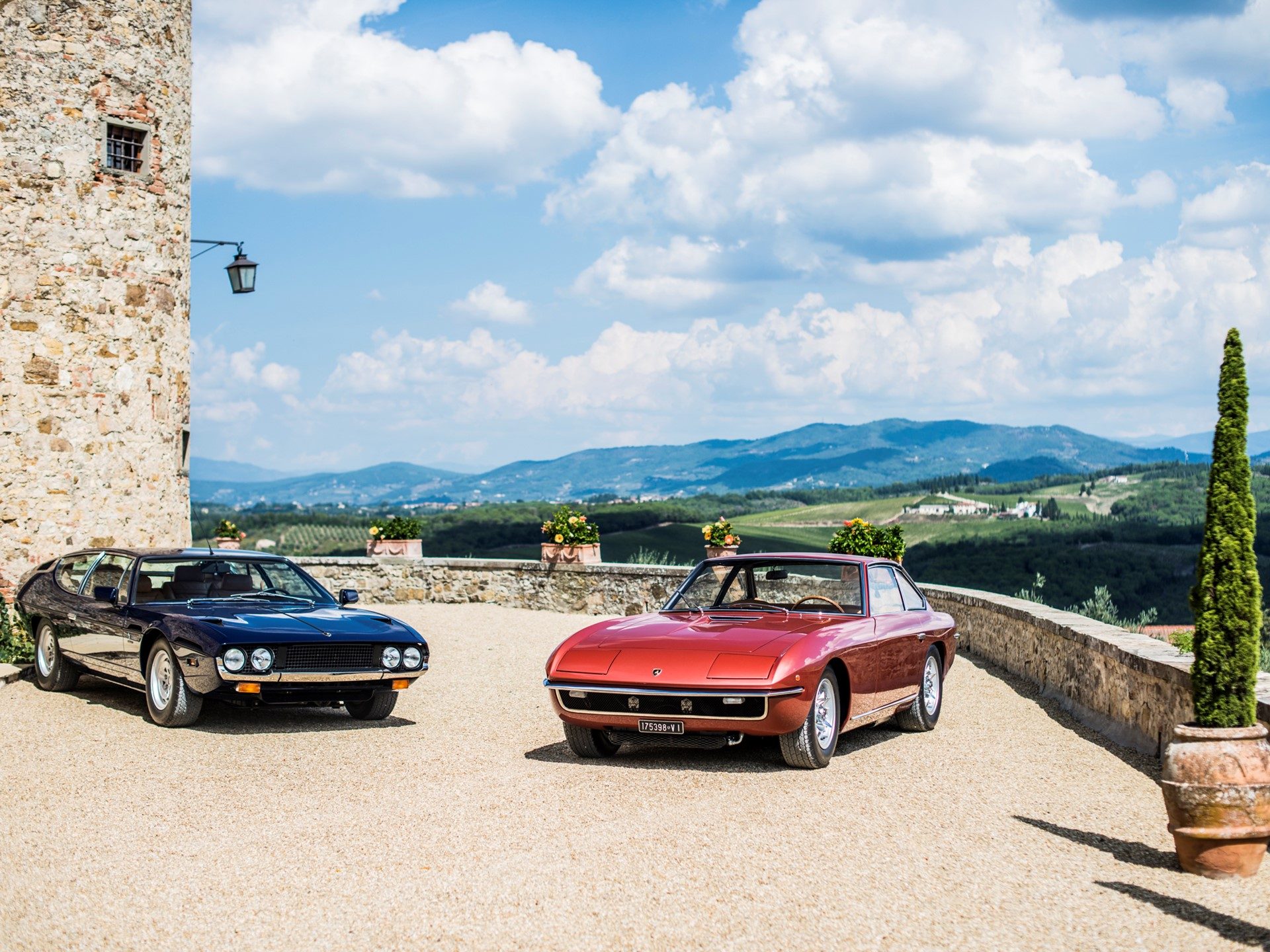 A dark blue Lamborghini Espada and a red Islero parked on a gravel driveway in front of a stone building in the Italian countryside