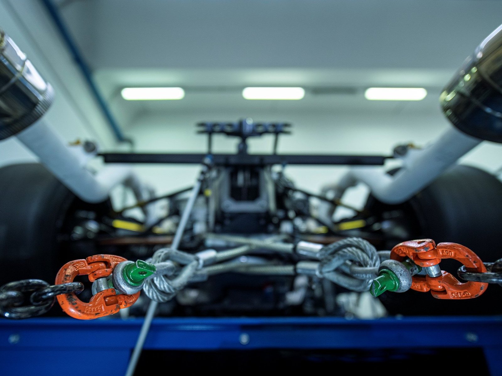 Close-up of heavy-duty orange shackles and steel cables with the Essenza SCV12's rear suspension and exhaust visible in a workshop