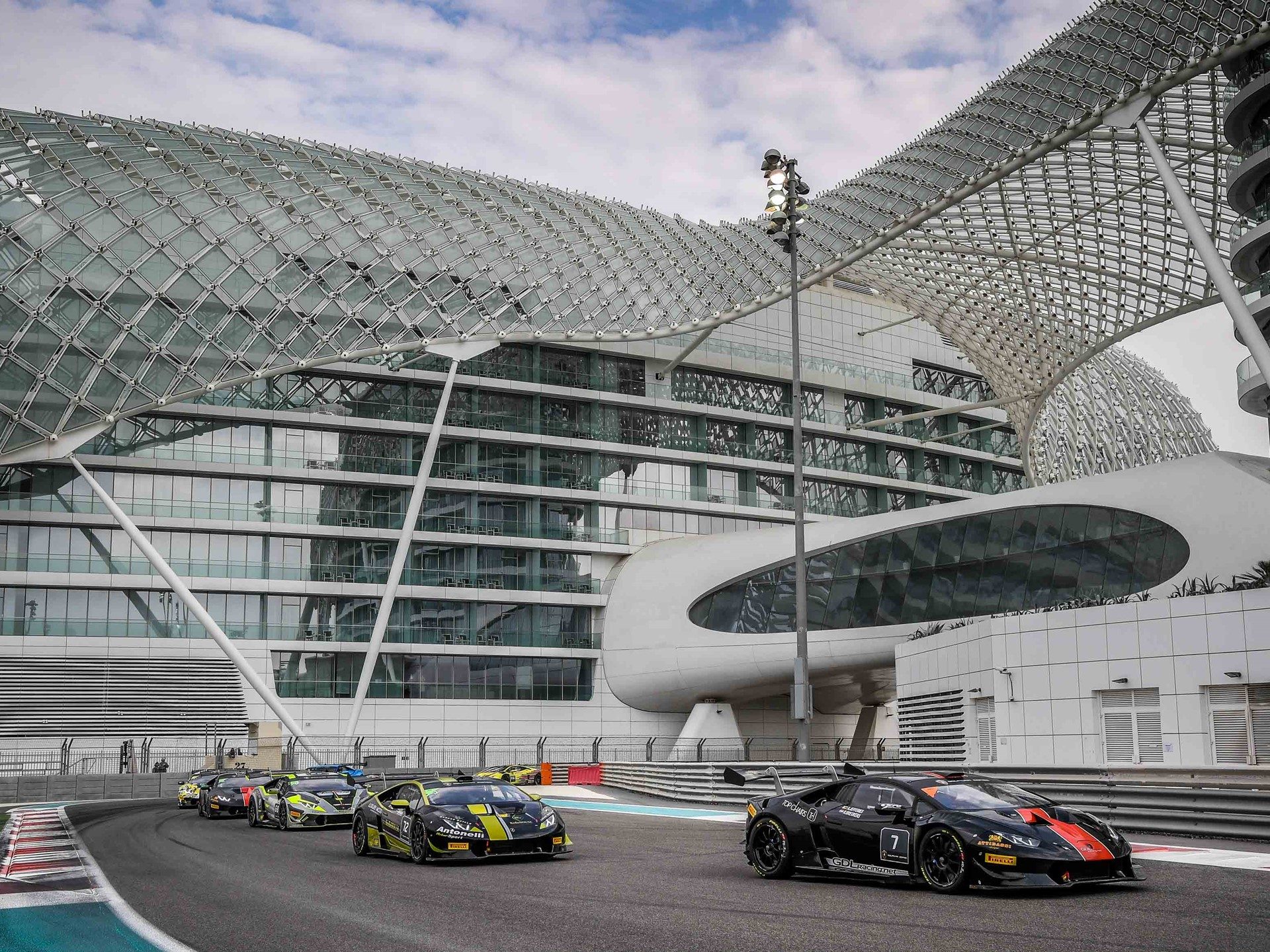 A pack of Lamborghini Huracán Super Trofeo race cars in black, orange, and yellow liveries navigate a corner at Yas Marina Circuit during the 2017 Super Trofeo Middle East round.