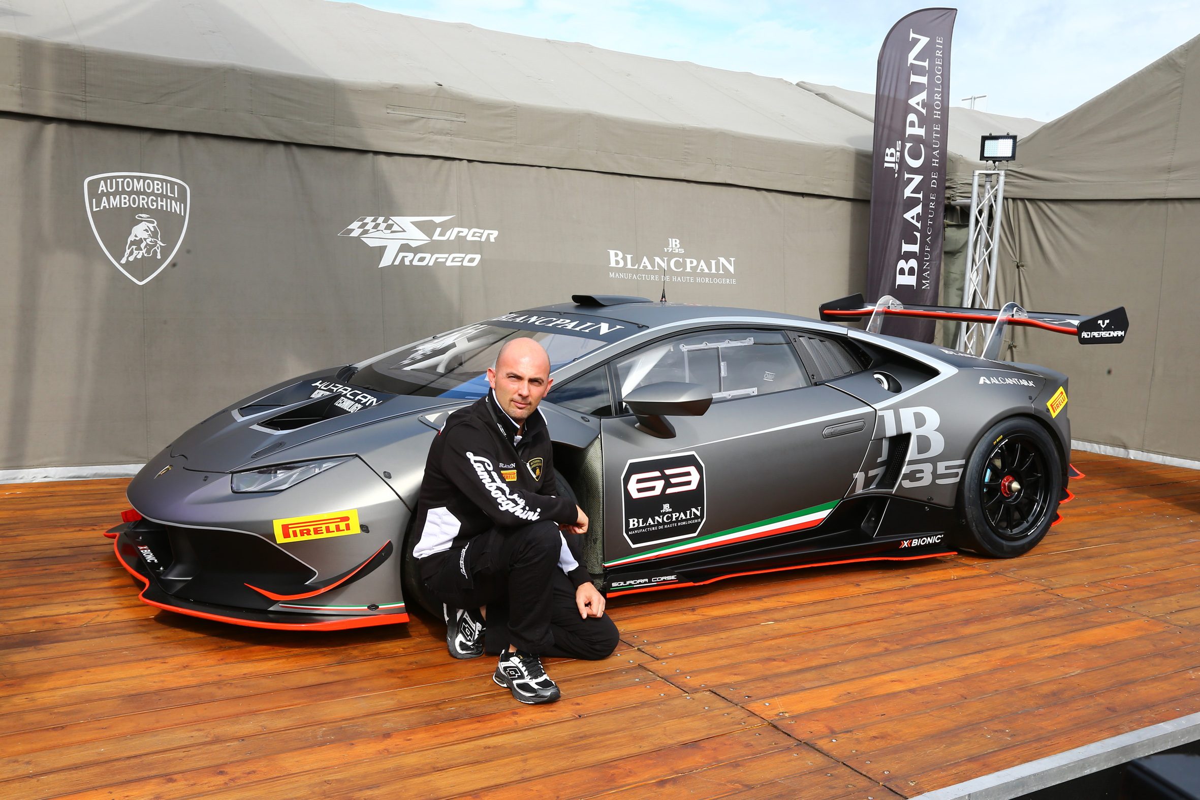 Giorgio Sanna kneeling beside a grey Lamborghini Huracán Super Trofeo race car at an outdoor event