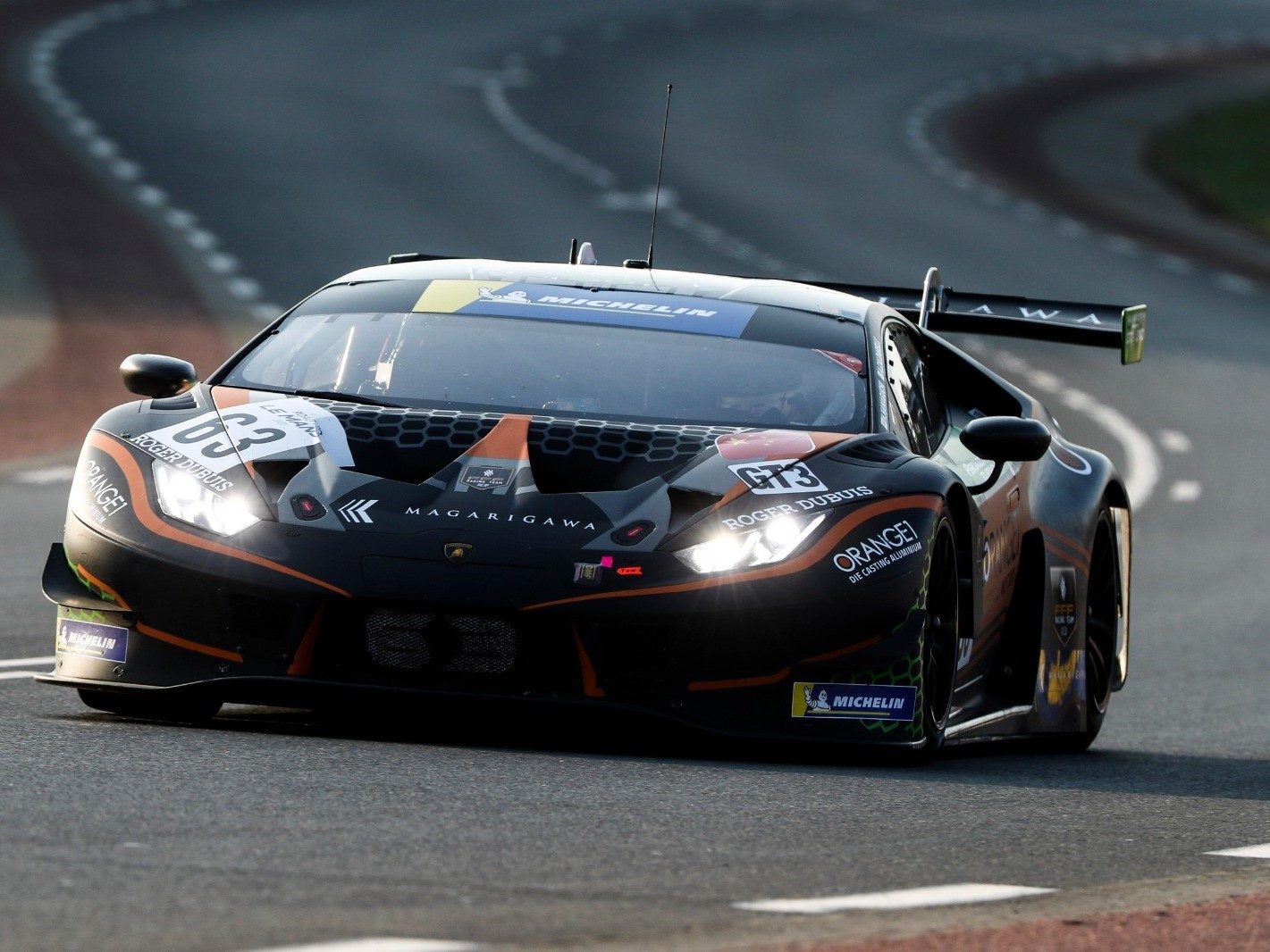 Black Lamborghini Huracán GT3 Evo race car with orange accents racing on track with headlights illuminated
