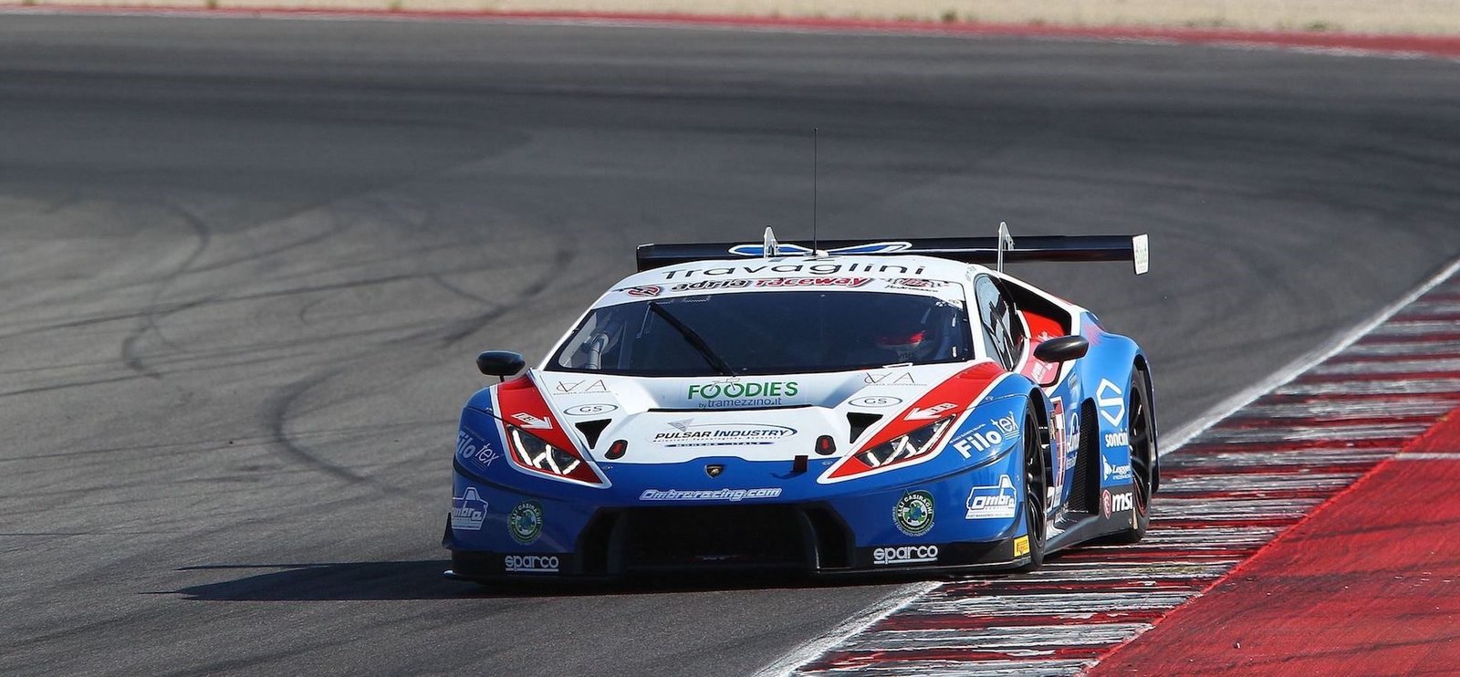 Blue, white, and red Lamborghini Huracán GT3 race car at speed on the Misano World Circuit, showcasing its large rear wing and racing livery
