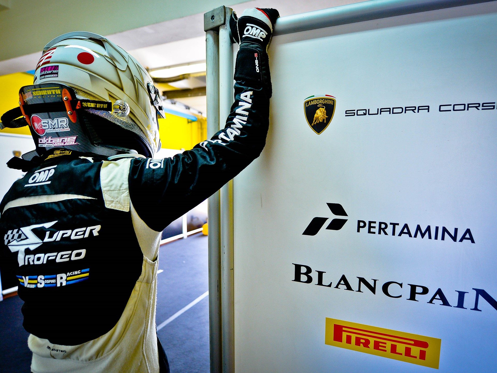 A racing driver in black and white racing suit leans against a wall bearing the Lamborghini Squadra Corse logo