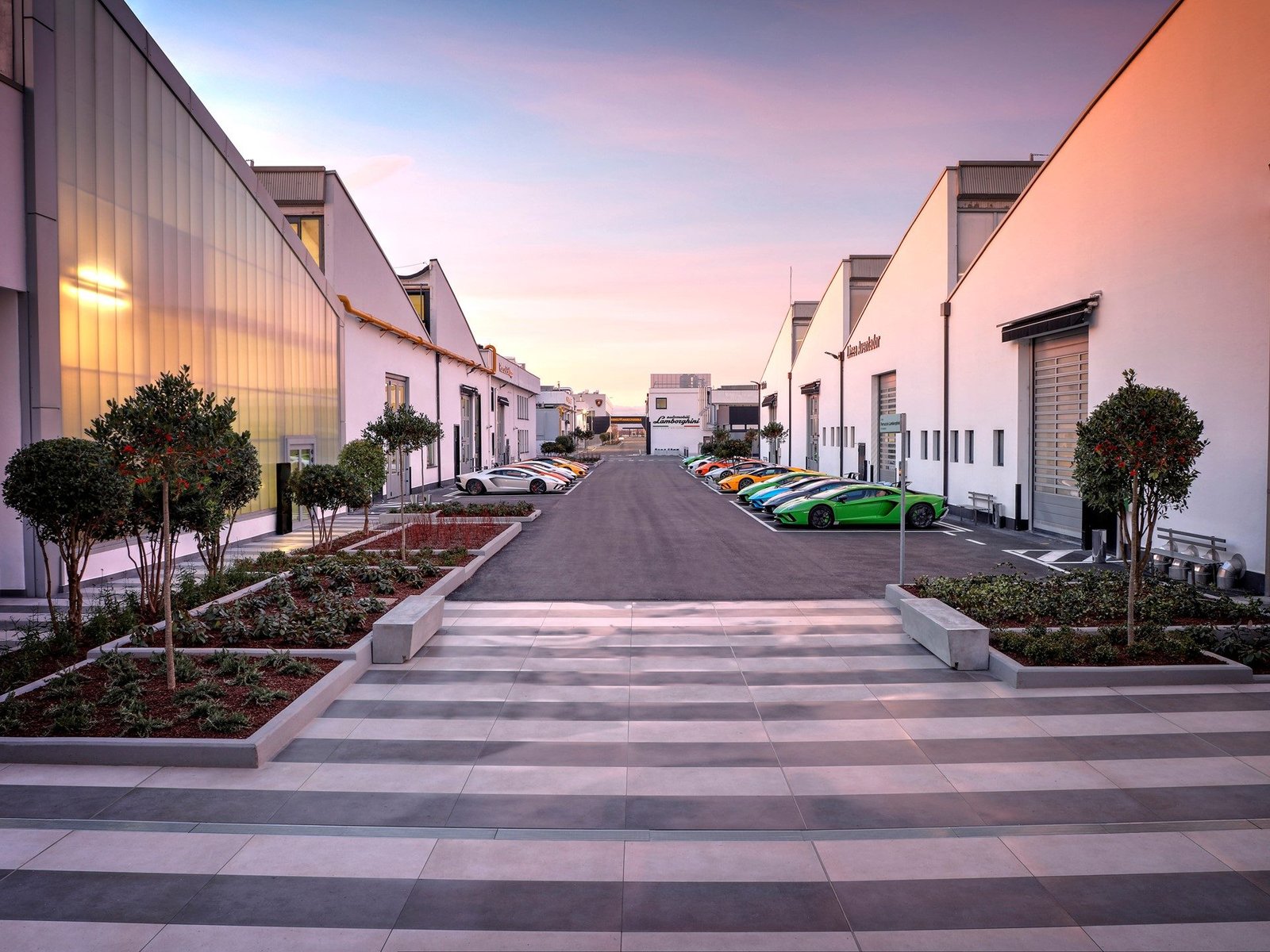 A lineup of colorful Lamborghini supercars parked along the factory road at Sant'Agata Bolognese at dusk, with modern white factory buildings on either side