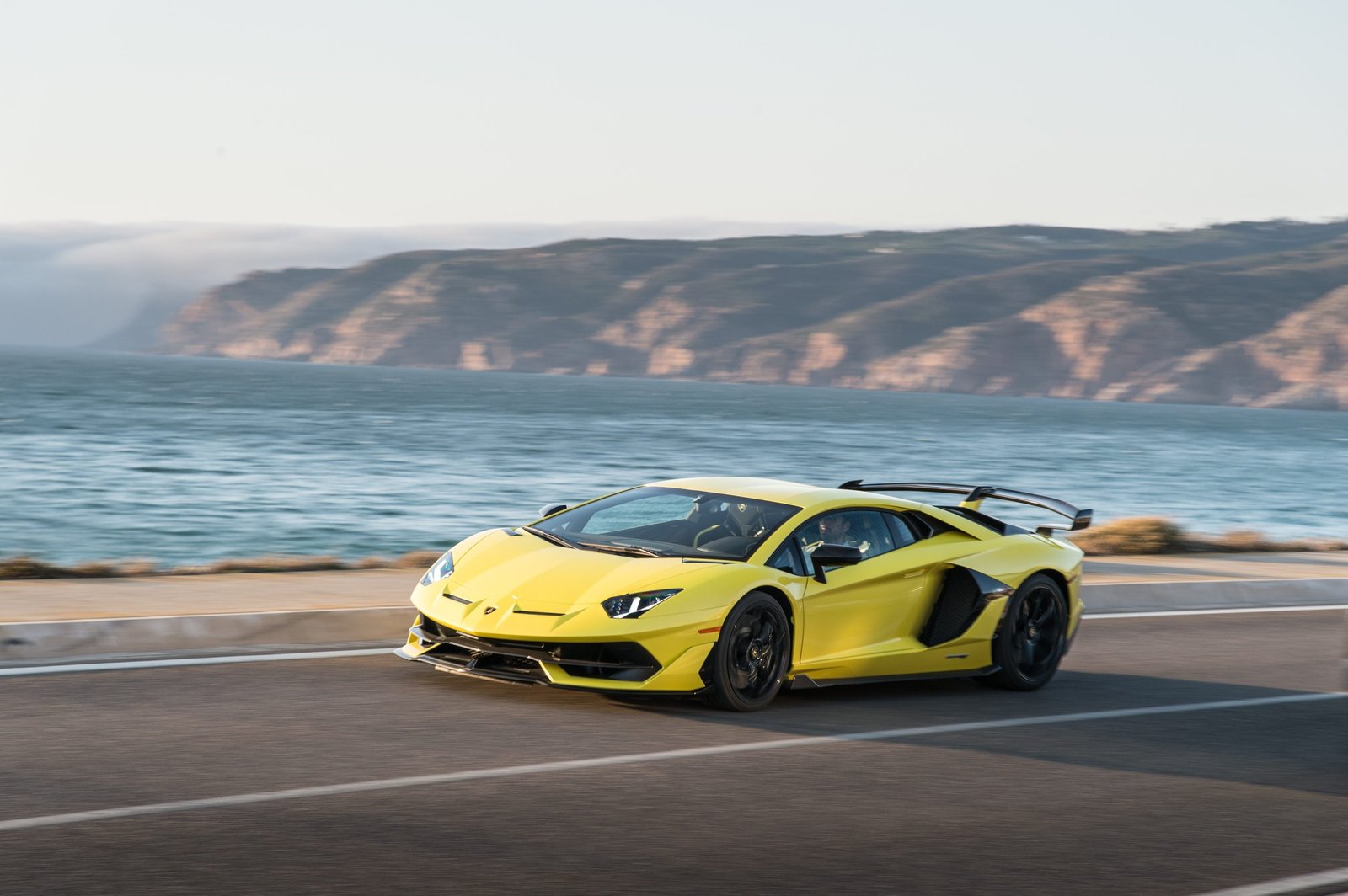 Yellow Lamborghini Aventador SVJ driving on a coastal road with ocean and cliffs in the background