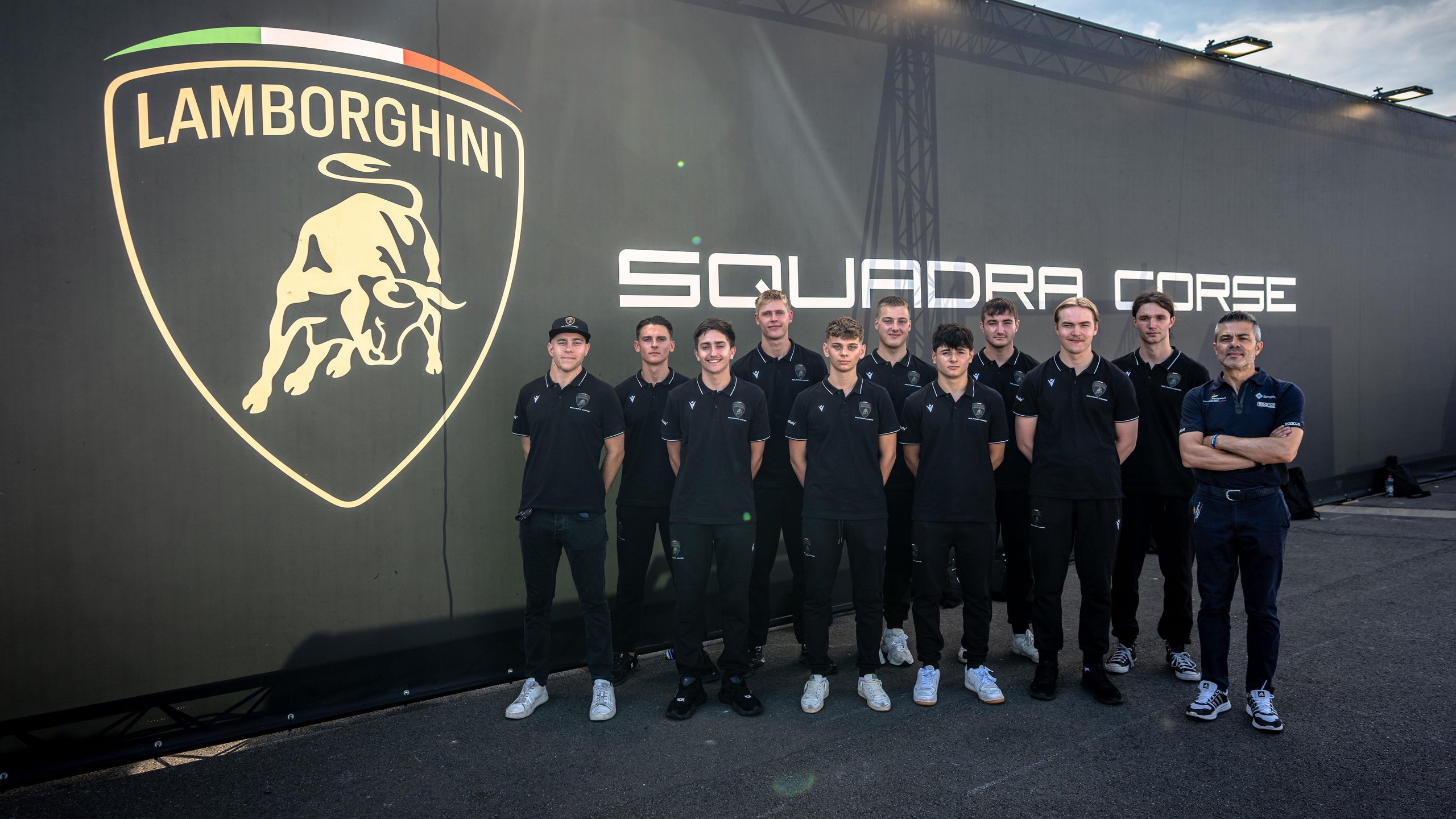 Thirty Drivers, One Goal: Inside Lamborghini'S 2026 Young Driver Programs And The Formula Medicine Gambit 3 Young drivers and a mentor standing in front of a lamborghini squadra corse banner