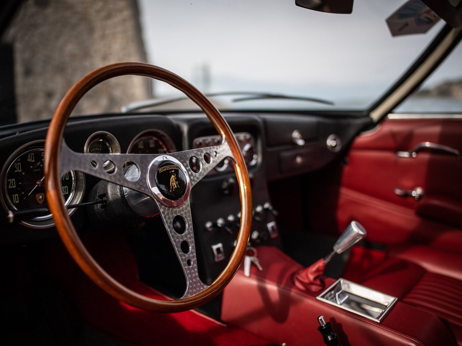 Interior of a classic Lamborghini grand tourer showing wooden steering wheel, chrome details, and red leather seats
