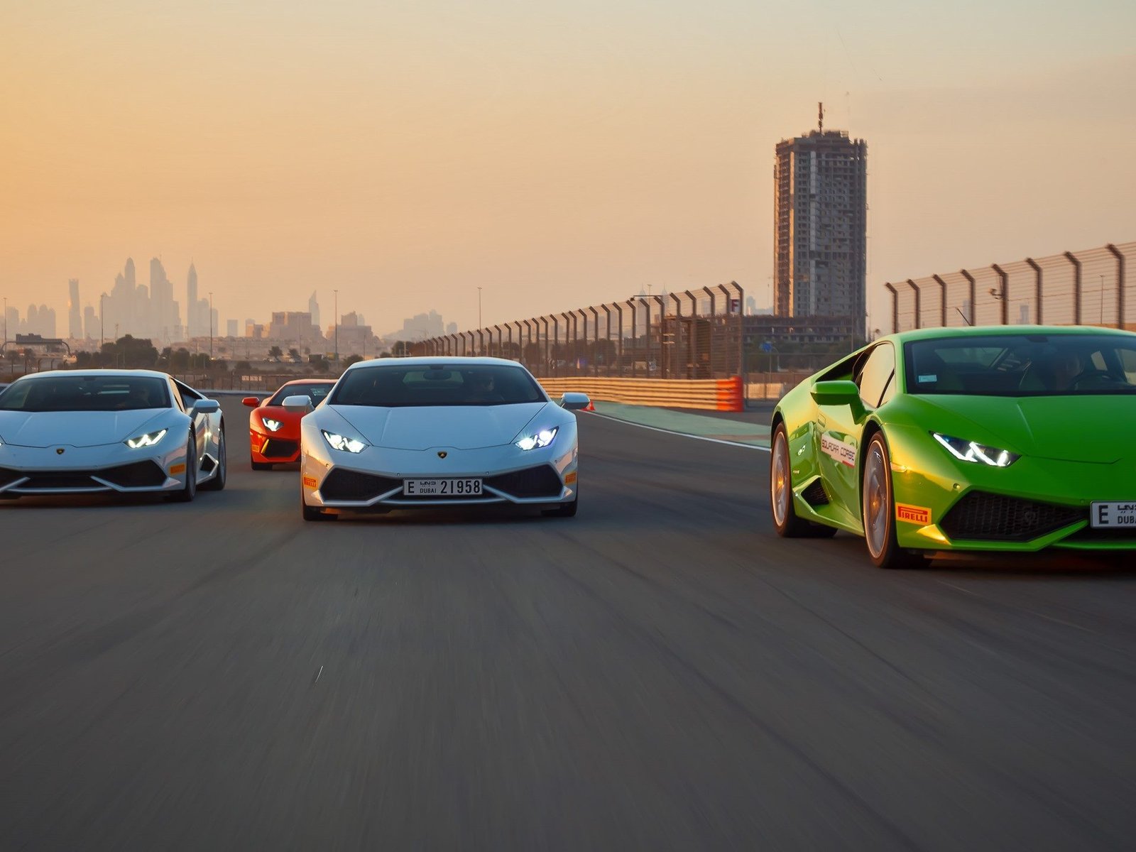 A fleet of Lamborghini Huracáns in green, white, and orange racing on a Dubai circuit at sunset