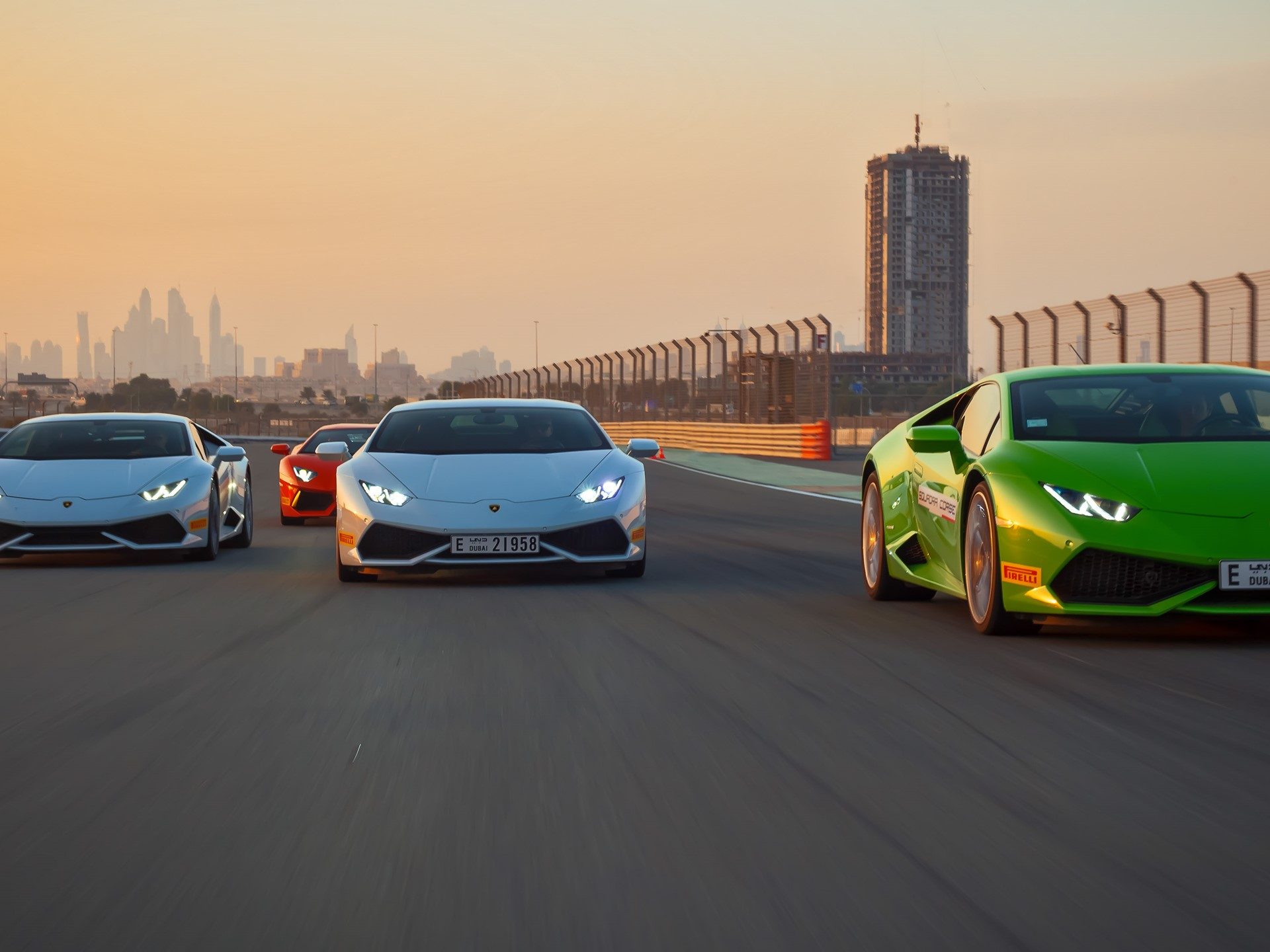 A fleet of Lamborghini Huracáns in green, white, and orange racing on a Dubai circuit at sunset