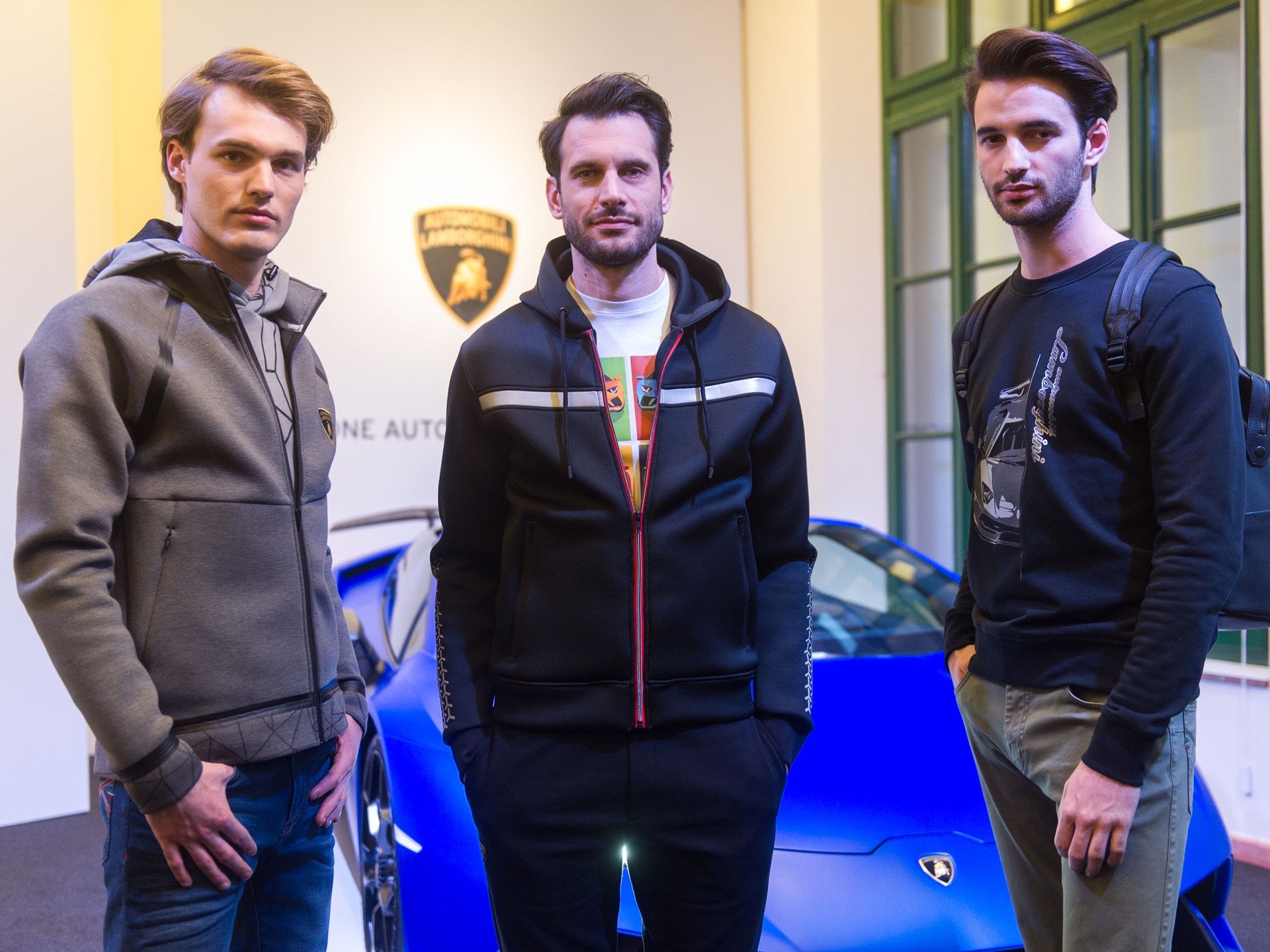 Three models in Lamborghini-branded casual apparel standing in front of a blue Huracán with the Lamborghini shield logo visible behind them