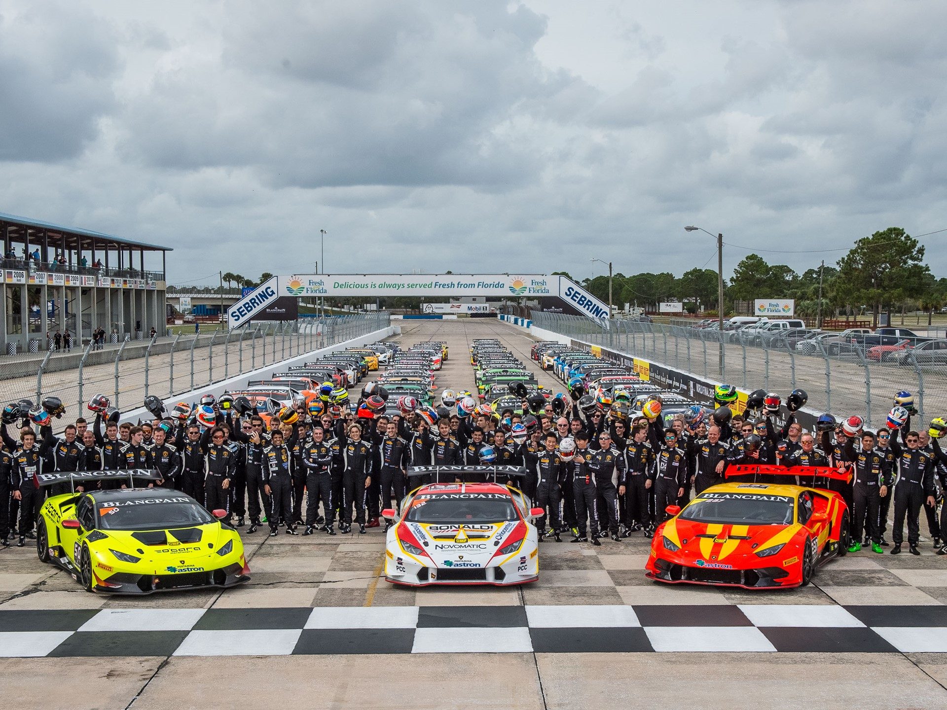 Three Lamborghini Huracán Super Trofeo race cars in yellow, white/red, and red/orange liveries on the grid with team members