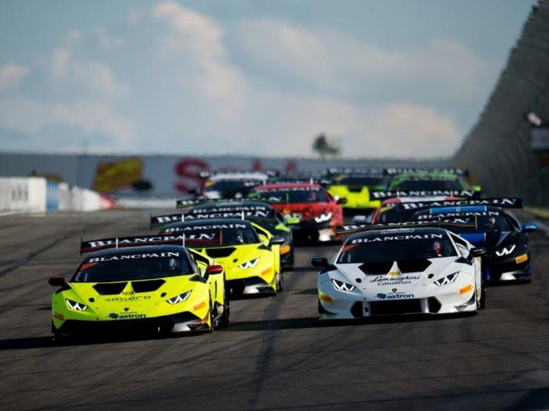 A pack of Lamborghini Huracán Super Trofeo race cars in yellow, white, and red liveries racing on a circuit under clear skies